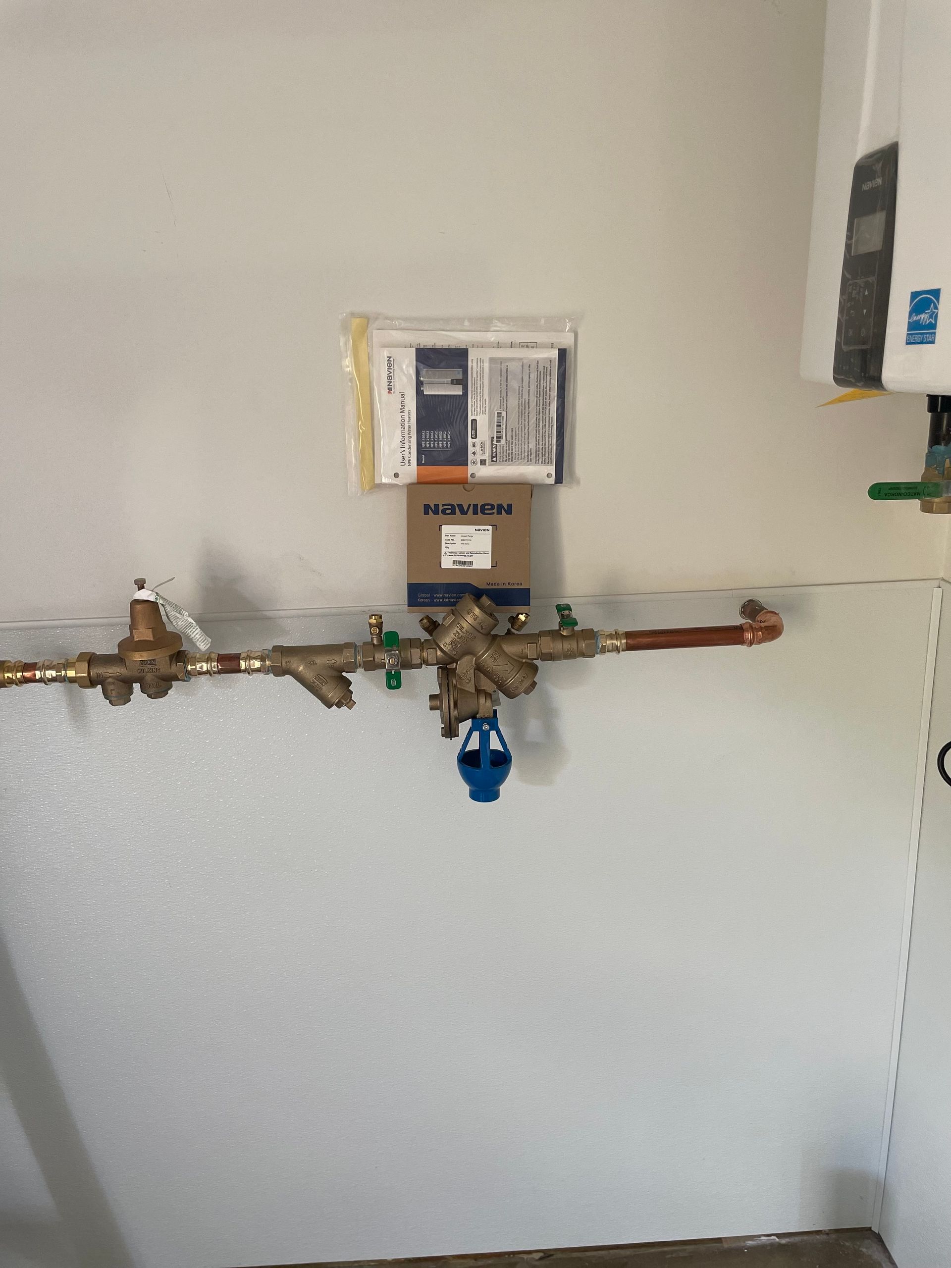 Water pipes with valves, including a blue backflow preventer, mounted on a white wall.