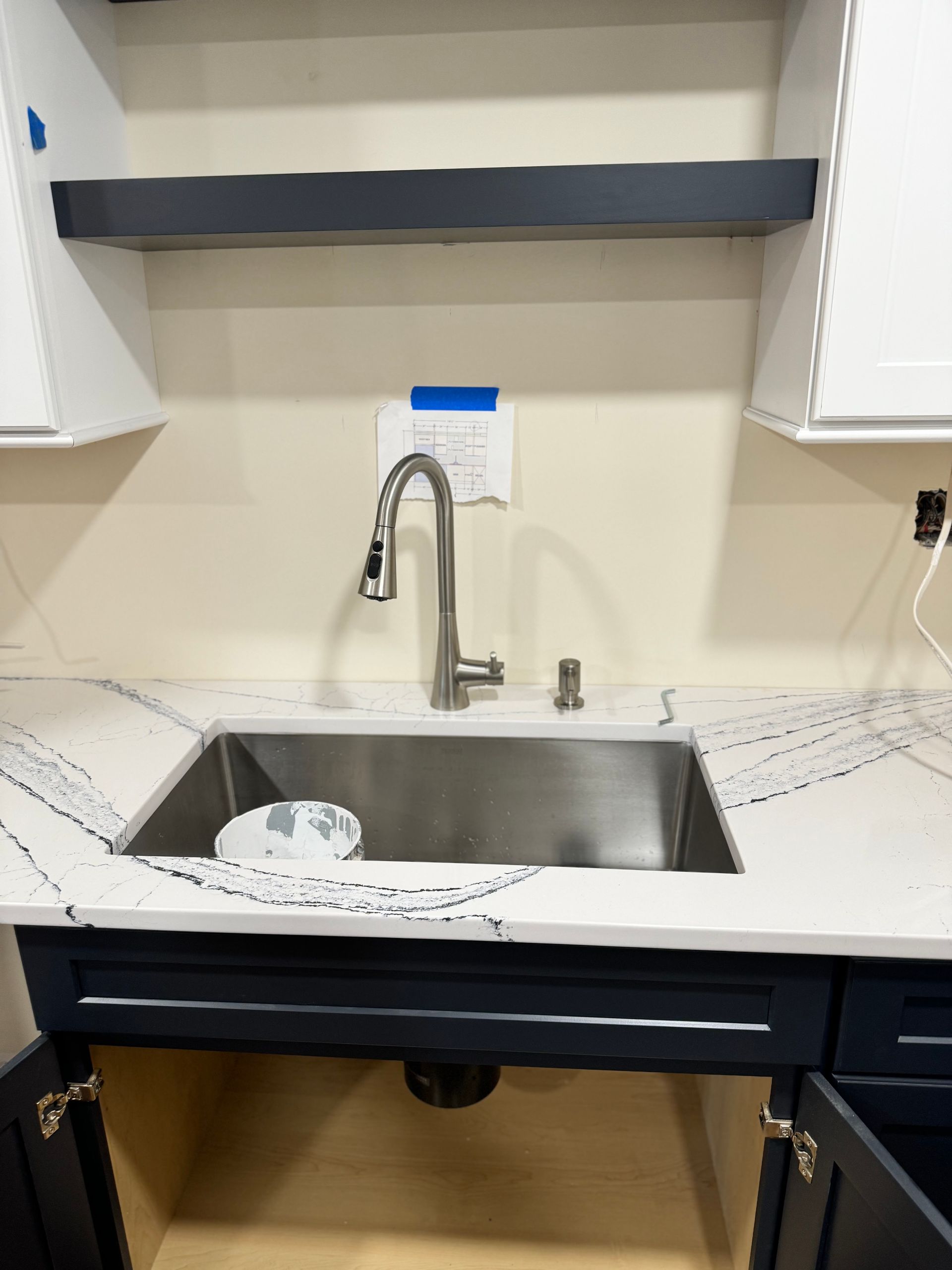 Kitchen sink area under construction, with dark blue cabinets, white countertops, and a stainless steel sink.