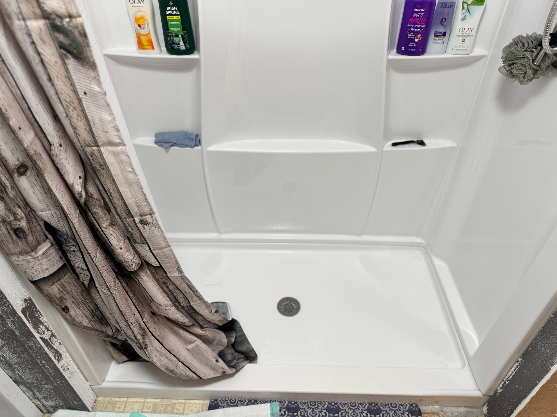 White shower stall with shower curtain, toiletries on shelves, and a drain.