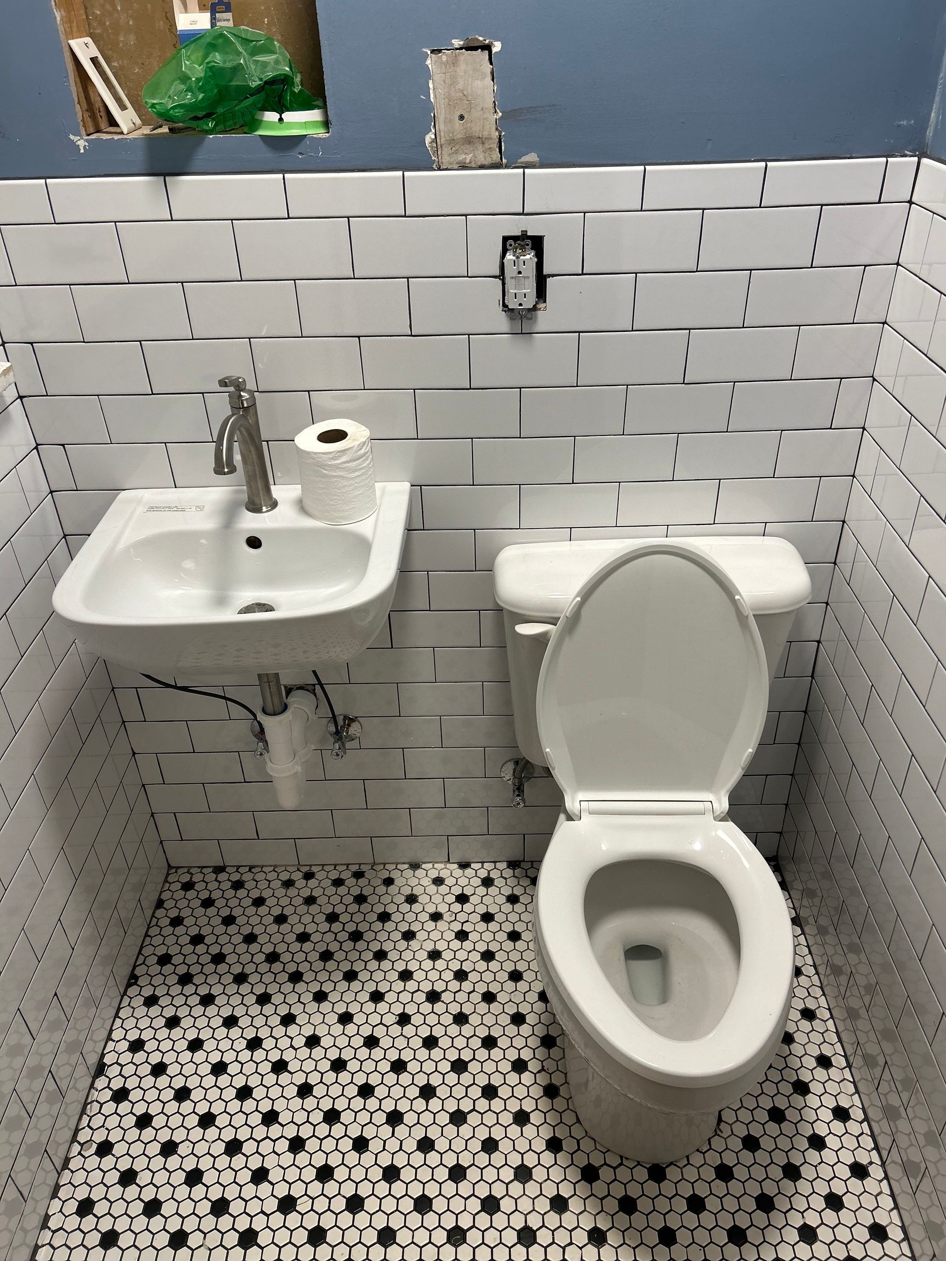 Small bathroom with white brick-like tiled walls, black and white patterned floor, toilet, sink, and toilet paper.