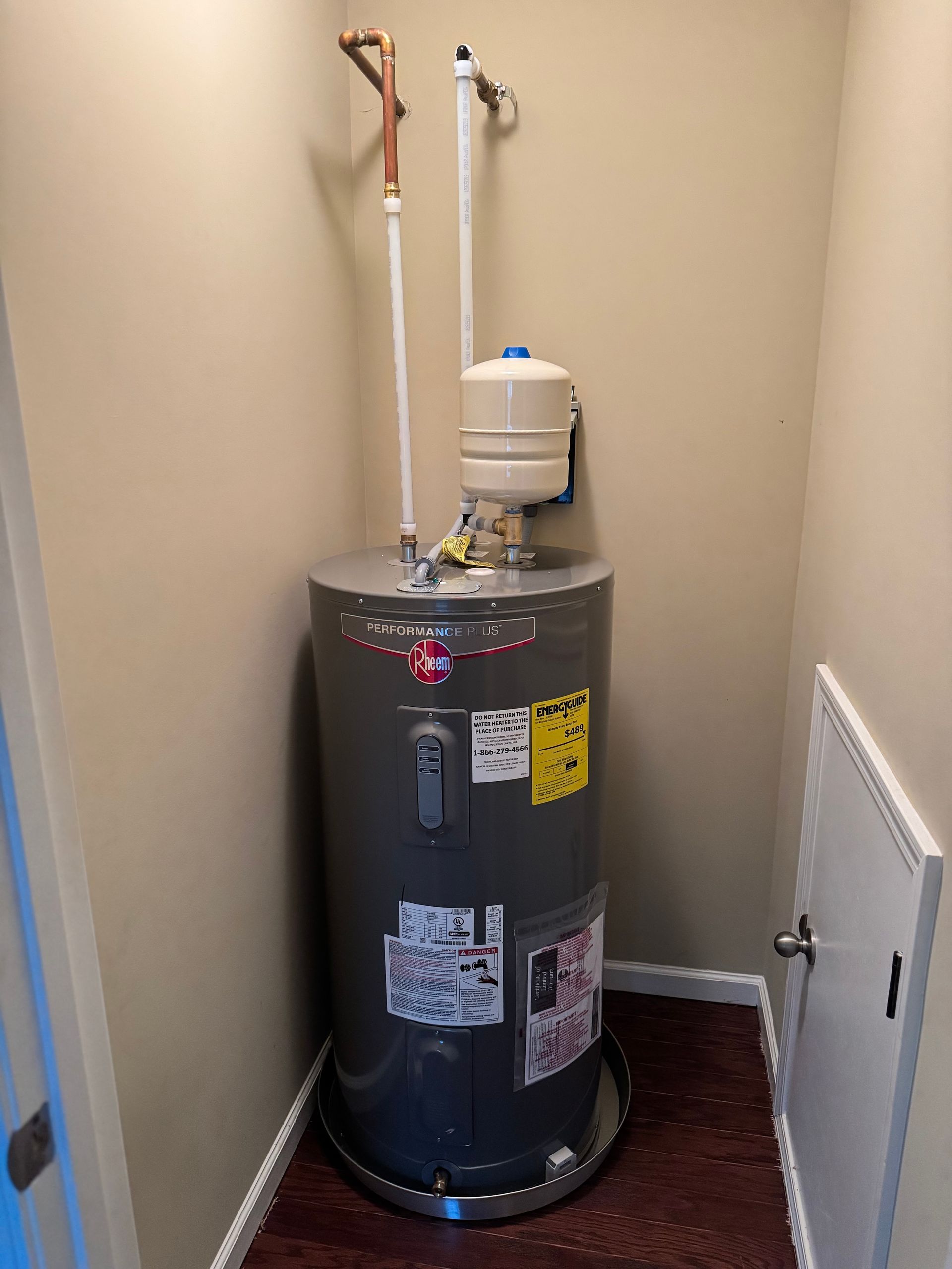 A tall gray water heater with white pipes in a small closet with a small access panel.
