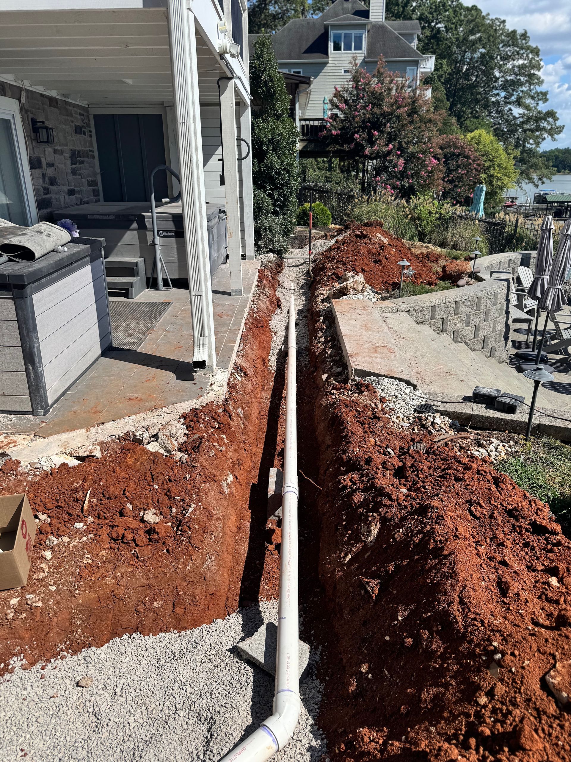 Trench dug alongside a house with a white pipe in it, surrounded by red soil and gravel.