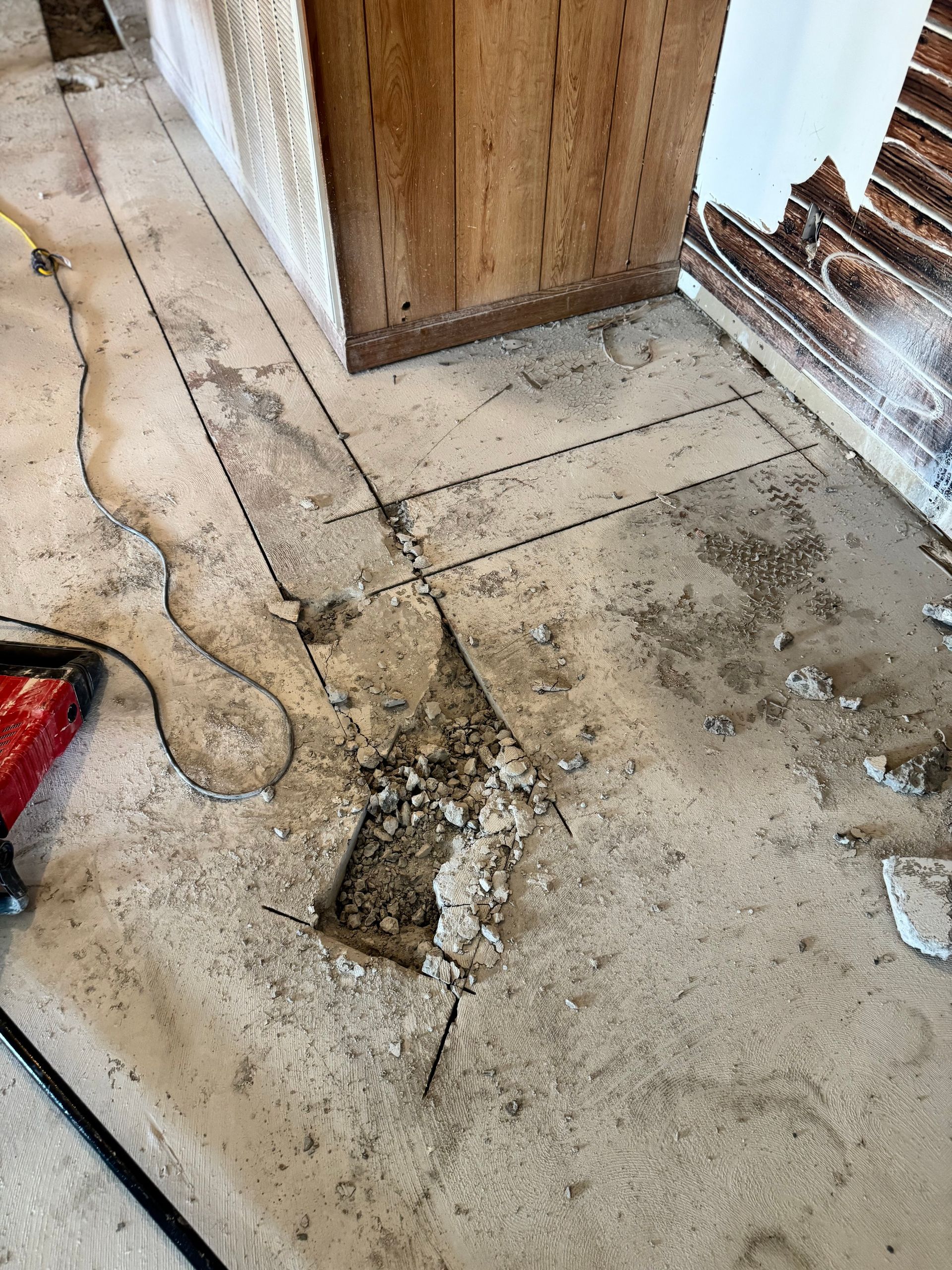 Damaged concrete floor in a kitchen with exposed subfloor. Sections marked for removal, tools present.