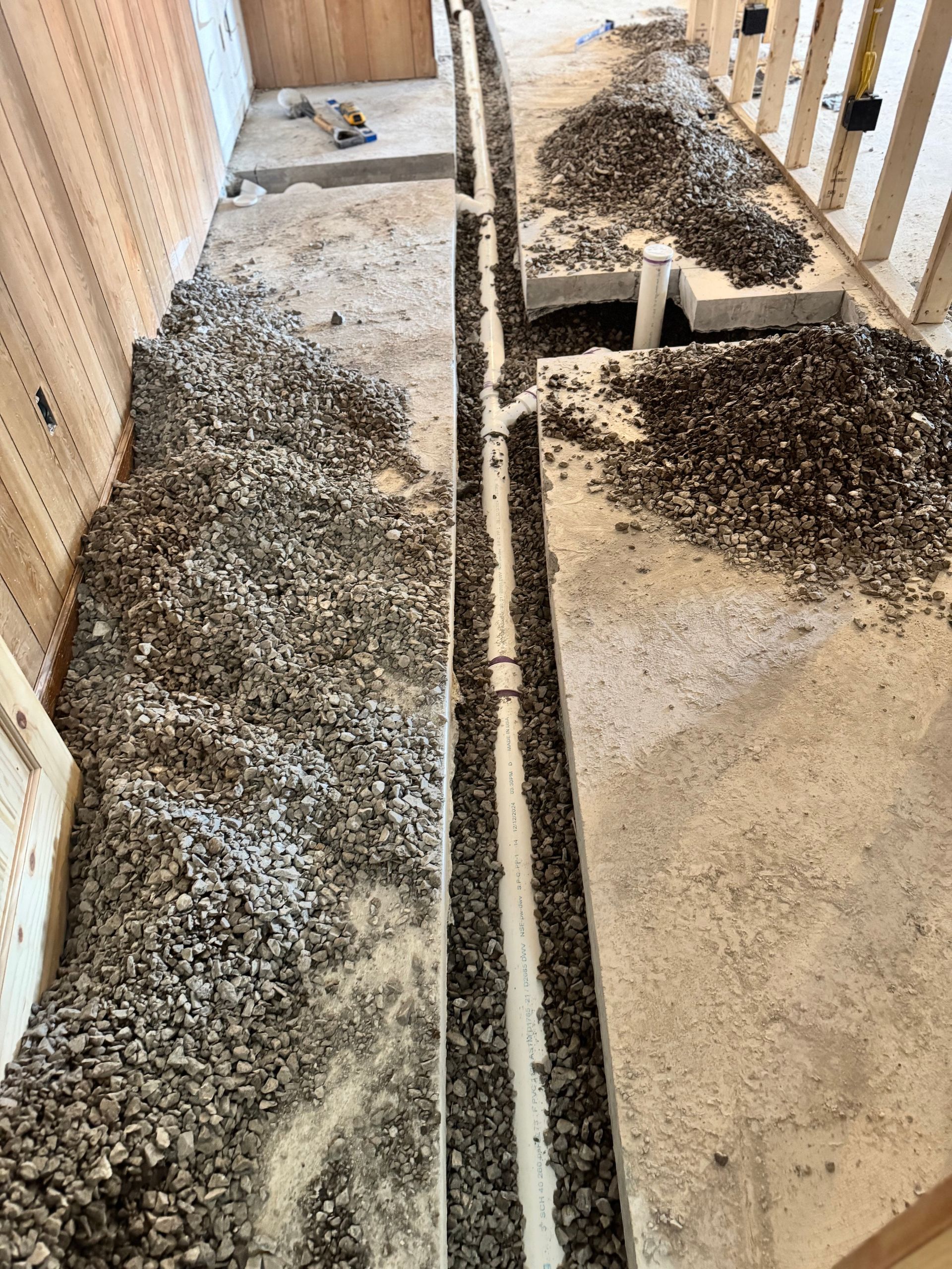 Construction site floor with trench for plumbing surrounded by gravel, unfinished walls visible.