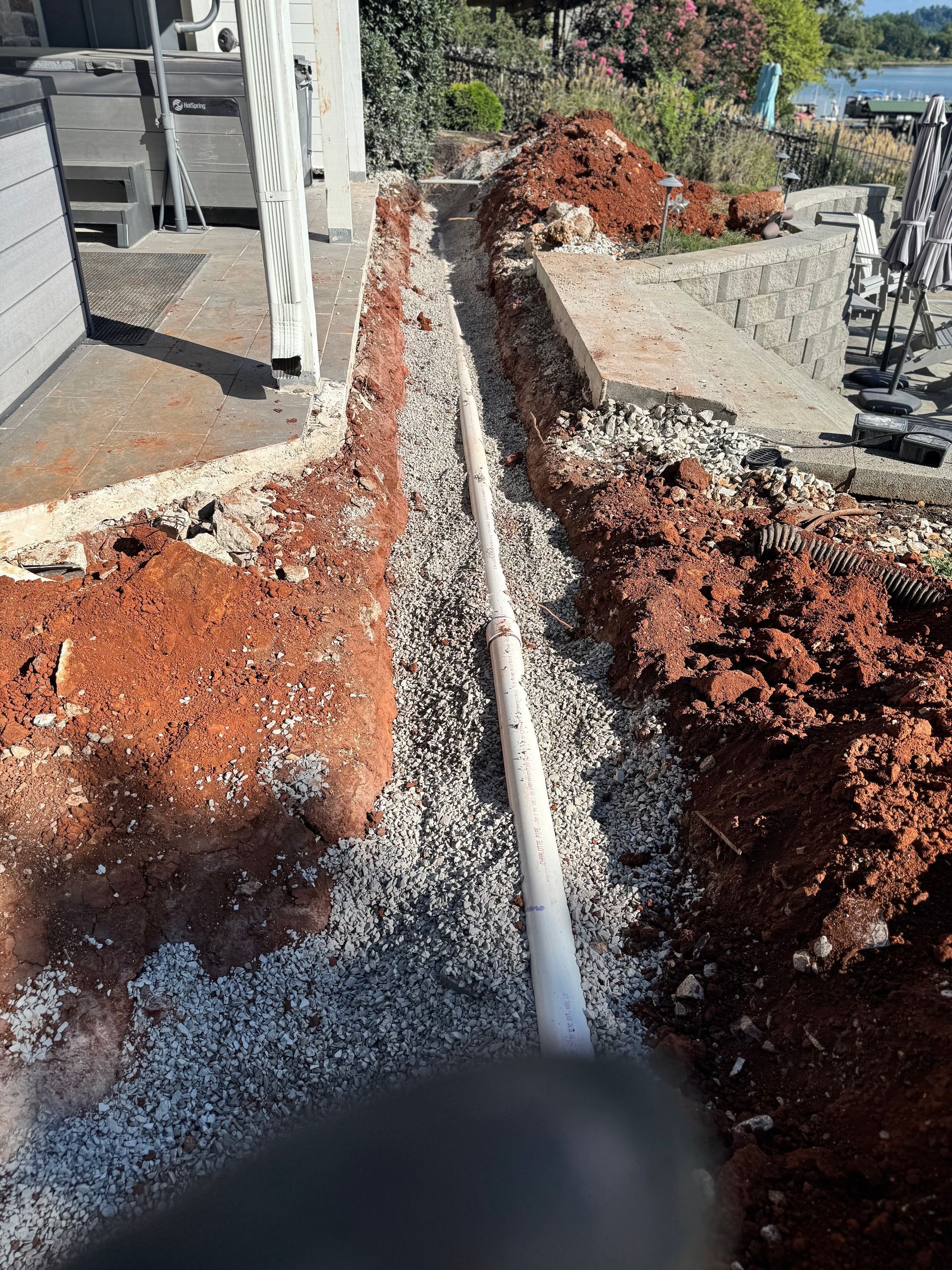 Trench with white pipe laid in gravel, surrounded by red soil, next to a building and retaining wall.