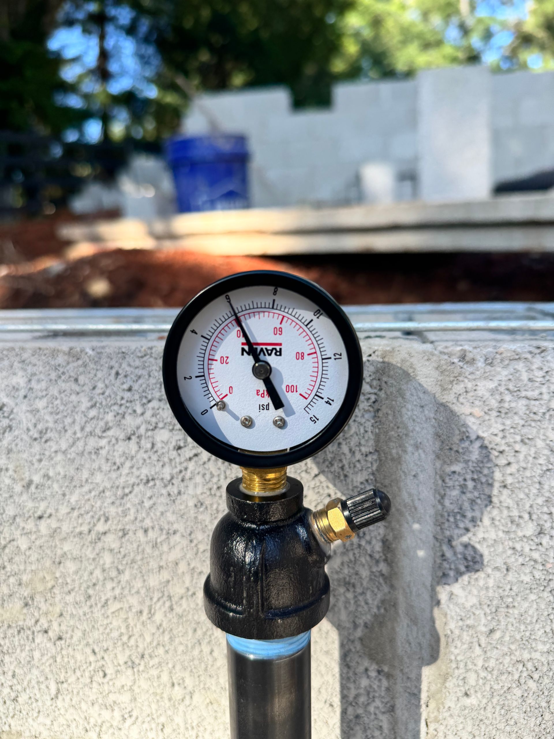 Pressure gauge attached to a pipe, reads approximately 4 PSI. Set against a concrete wall backdrop.