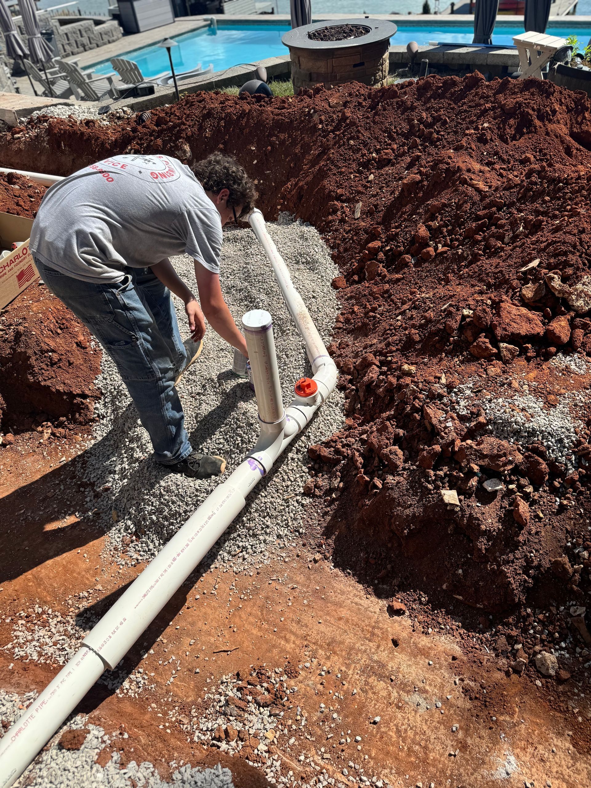 Man working with PVC pipes in a dirt trench, likely for landscaping or pool construction near a pool.