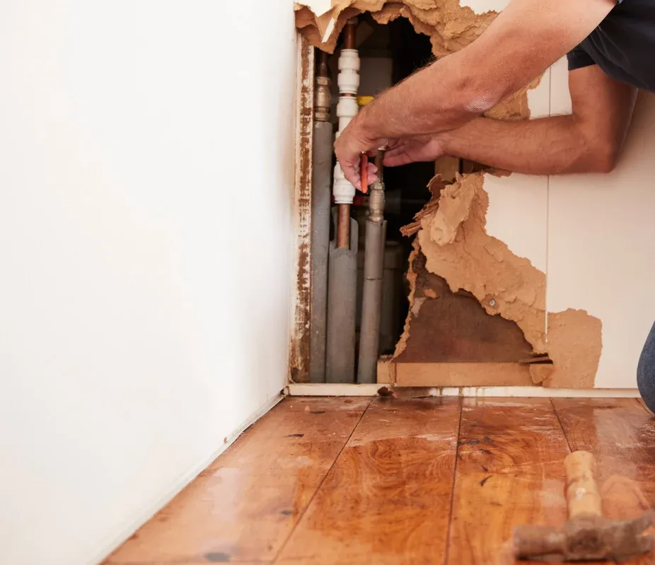 A person repairing pipes in a wall, revealing damaged drywall and wooden flooring.