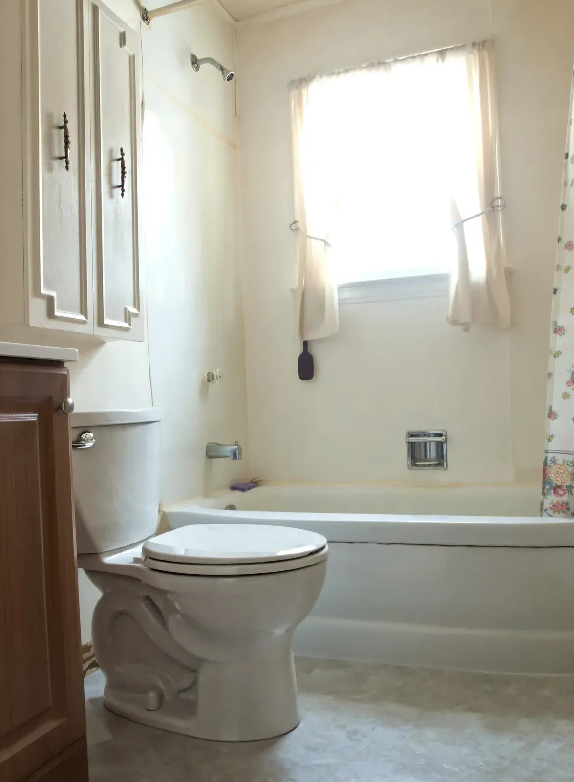 Bathroom with toilet, tub, cabinets, and a window with sheer curtains.