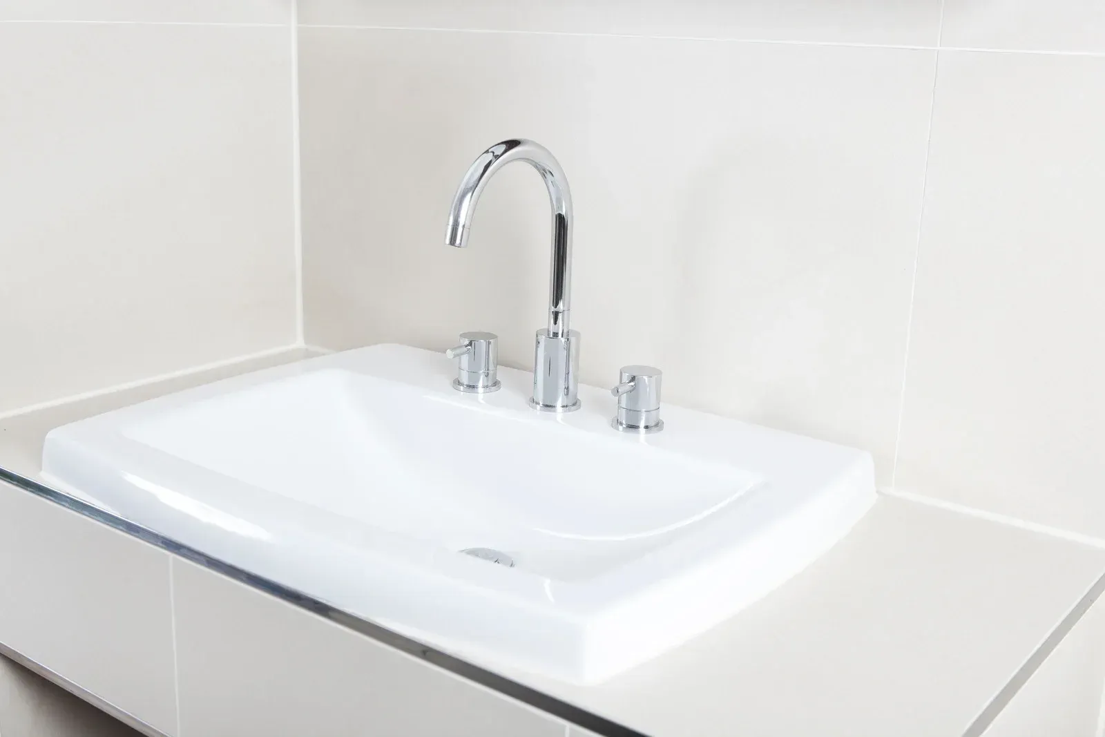 White sink with chrome faucet and knobs in a beige bathroom.