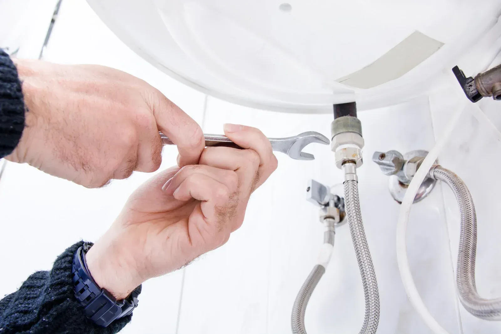 Hands using a wrench to tighten a pipe connection to a white water heater in a bathroom.