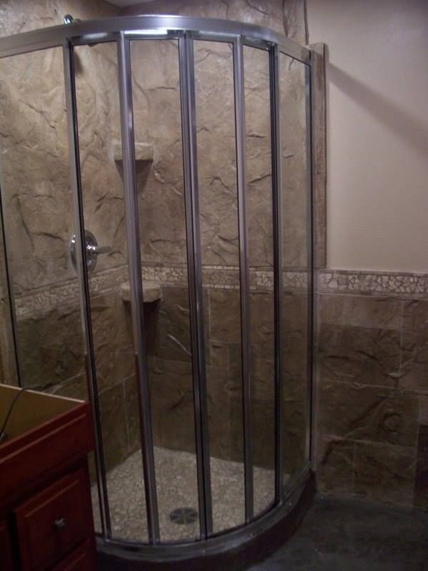 Curved shower enclosure with stone tile walls and a small, brown vanity on the left.
