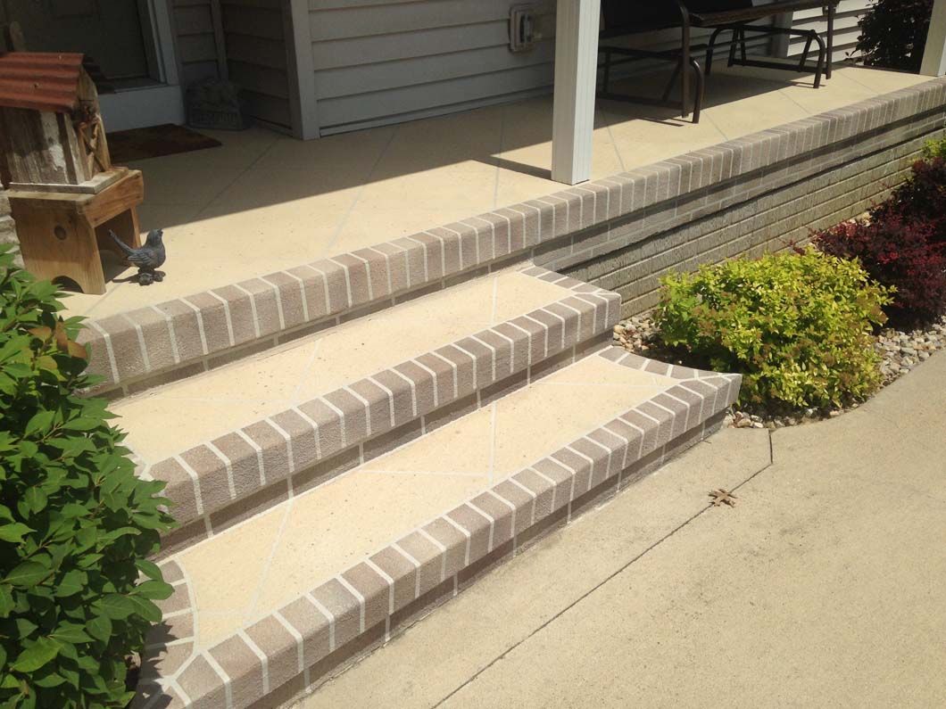 Brick steps leading up to a porch, with beige concrete and greenery on either side.