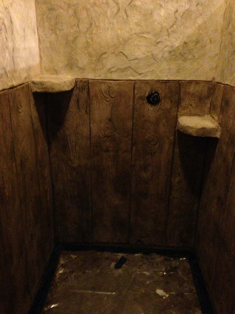 Small, dark shower with wood-paneled walls and stone shelves. The floor is dirty, and the walls are textured.