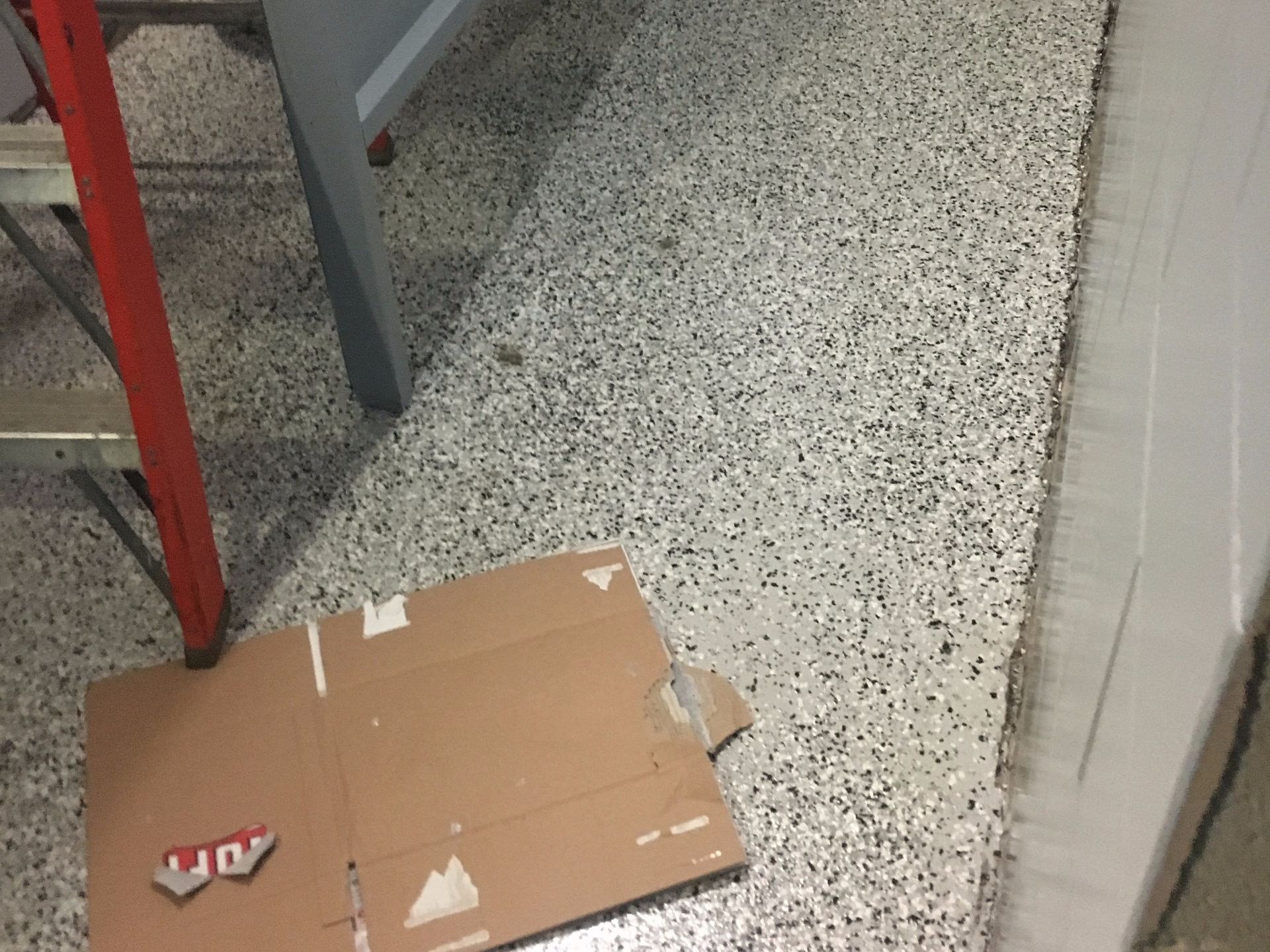 Cardboard on a speckled floor by a ladder and cabinets in a work area.