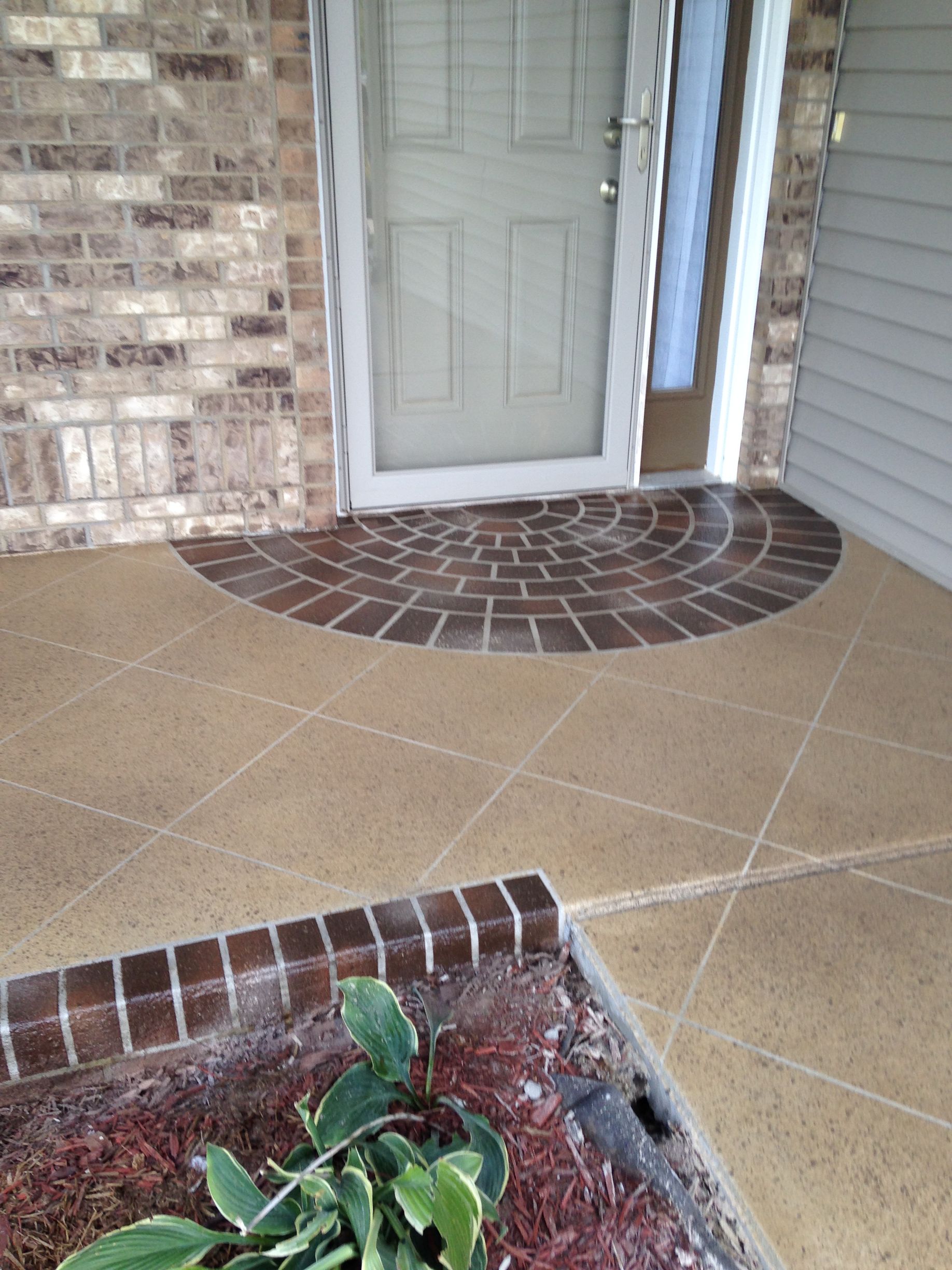 Entryway with brick and brown concrete, front door, and a small garden.