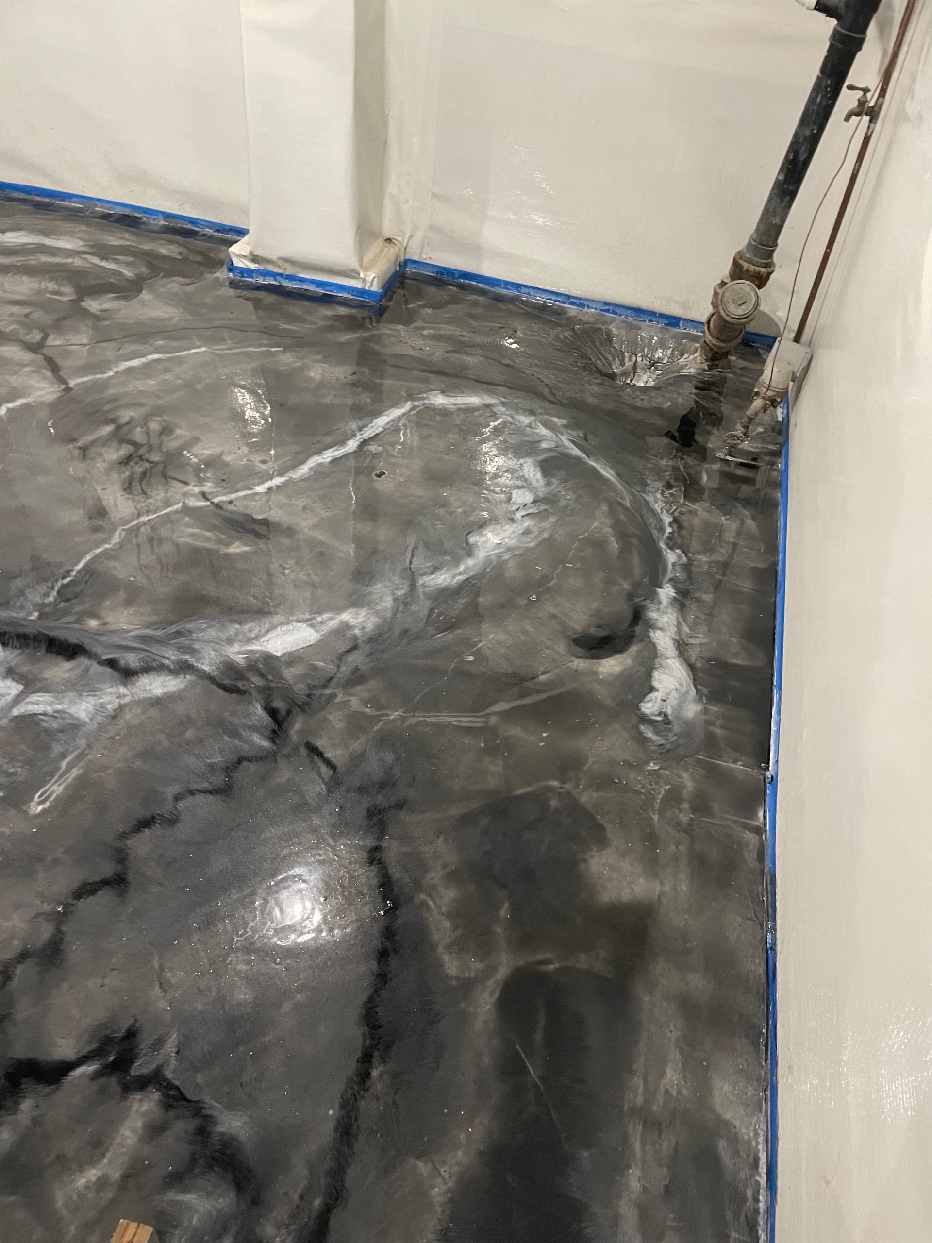 Grey and white swirled epoxy floor with cracks, blue tape along edges, near pipes and white wall.