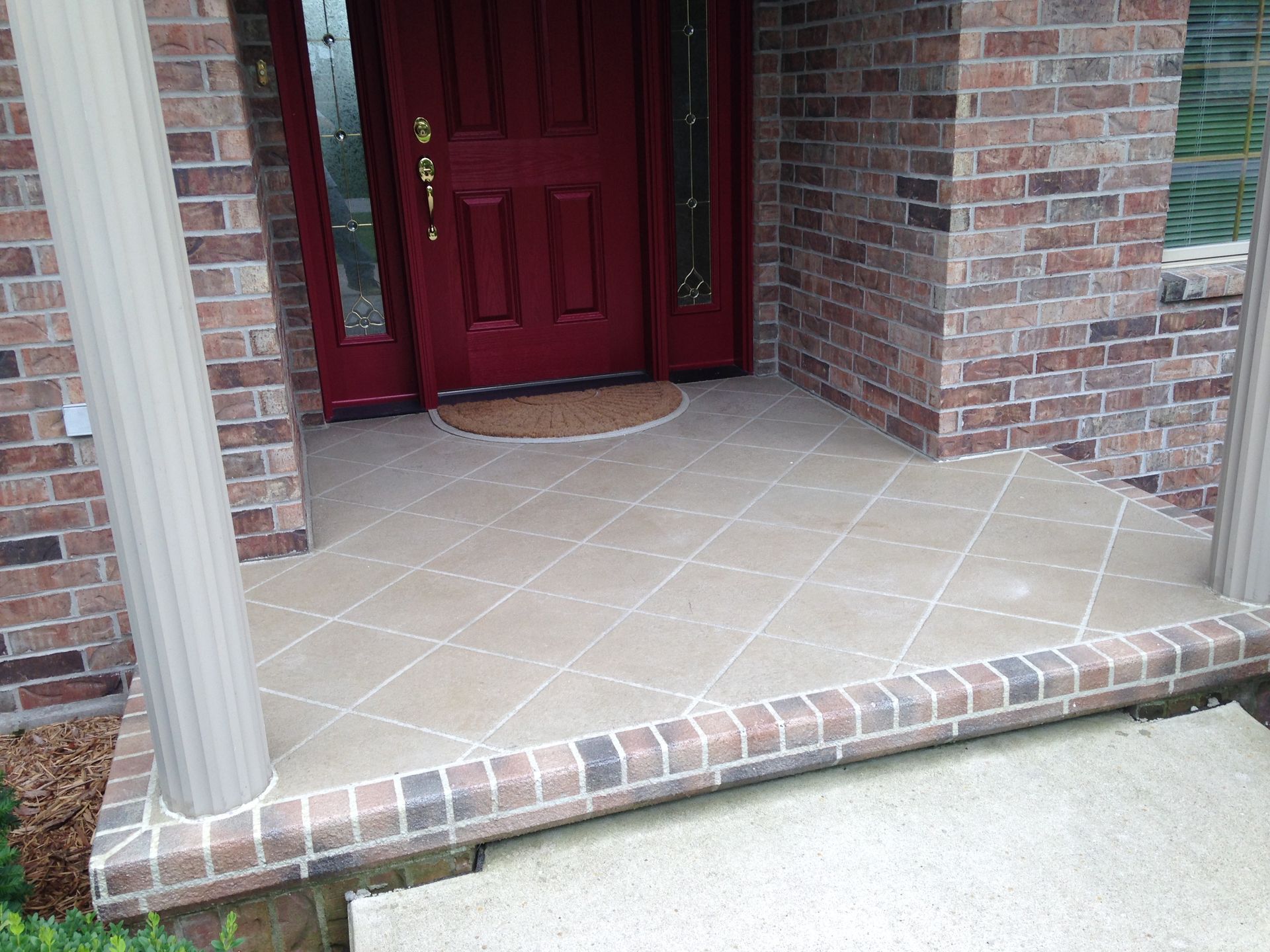 Brick porch with red door, tan floor with diagonal lines, bordered by bricks, and white columns.