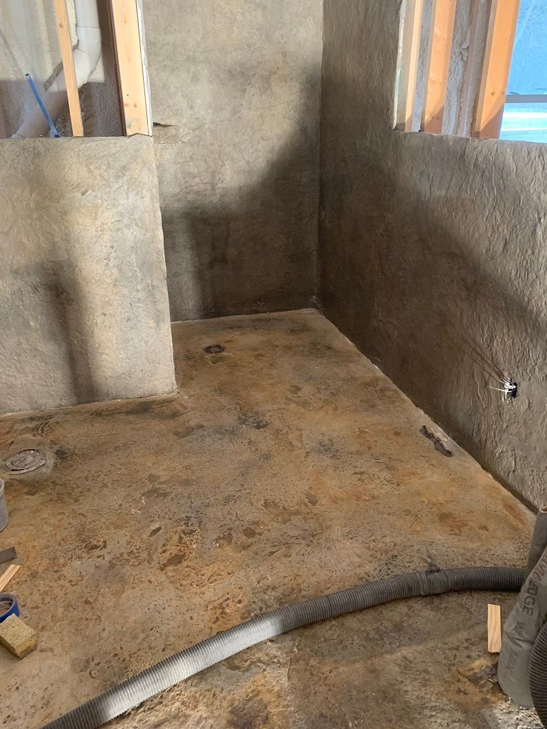 A small room under construction with gray walls and a concrete floor. A dark hose lies across the floor.