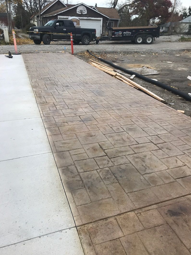 Stamped concrete driveway, with dark brown paving, next to a gray concrete slab. A truck and trailer are in the background.