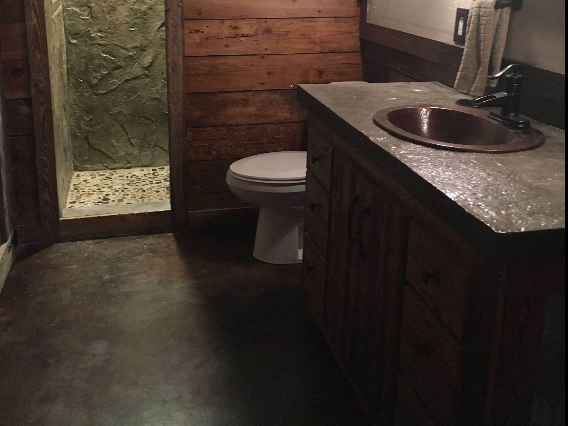 Rustic bathroom with wooden walls, concrete floor, and stone-tiled shower. Copper sink.