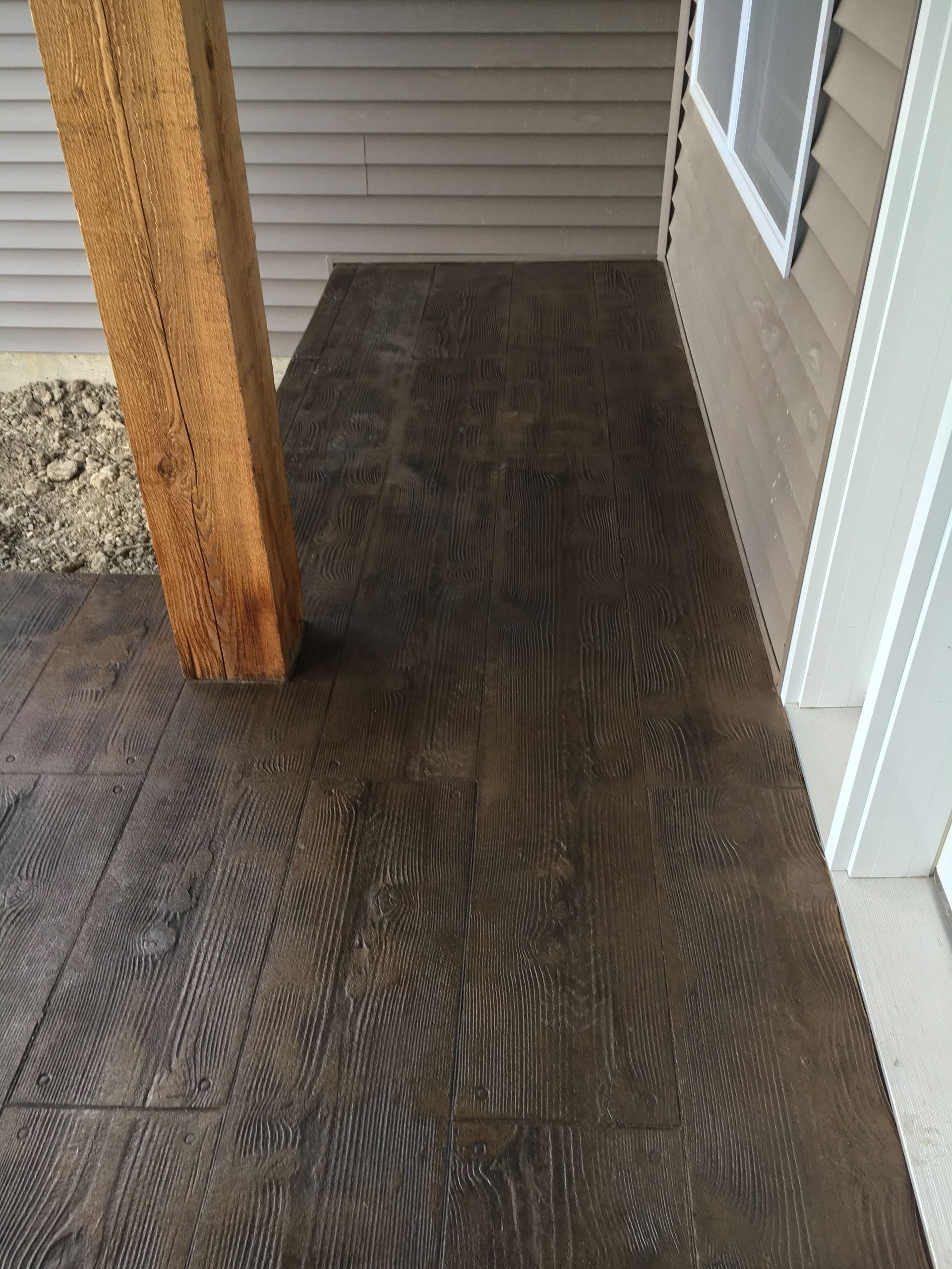 Dark brown wood-grain stamped concrete porch, tan siding, wooden post, and window frame.