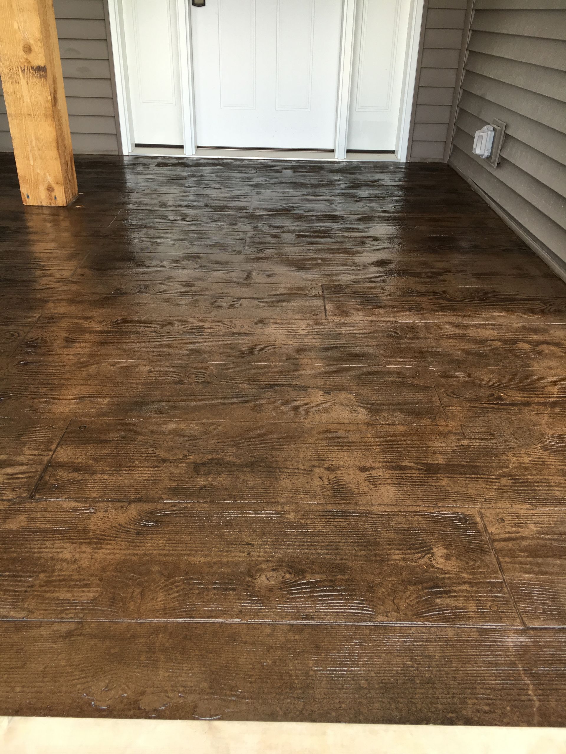 Brown wood-grain stamped concrete porch floor leading to a white door. The porch appears wet.