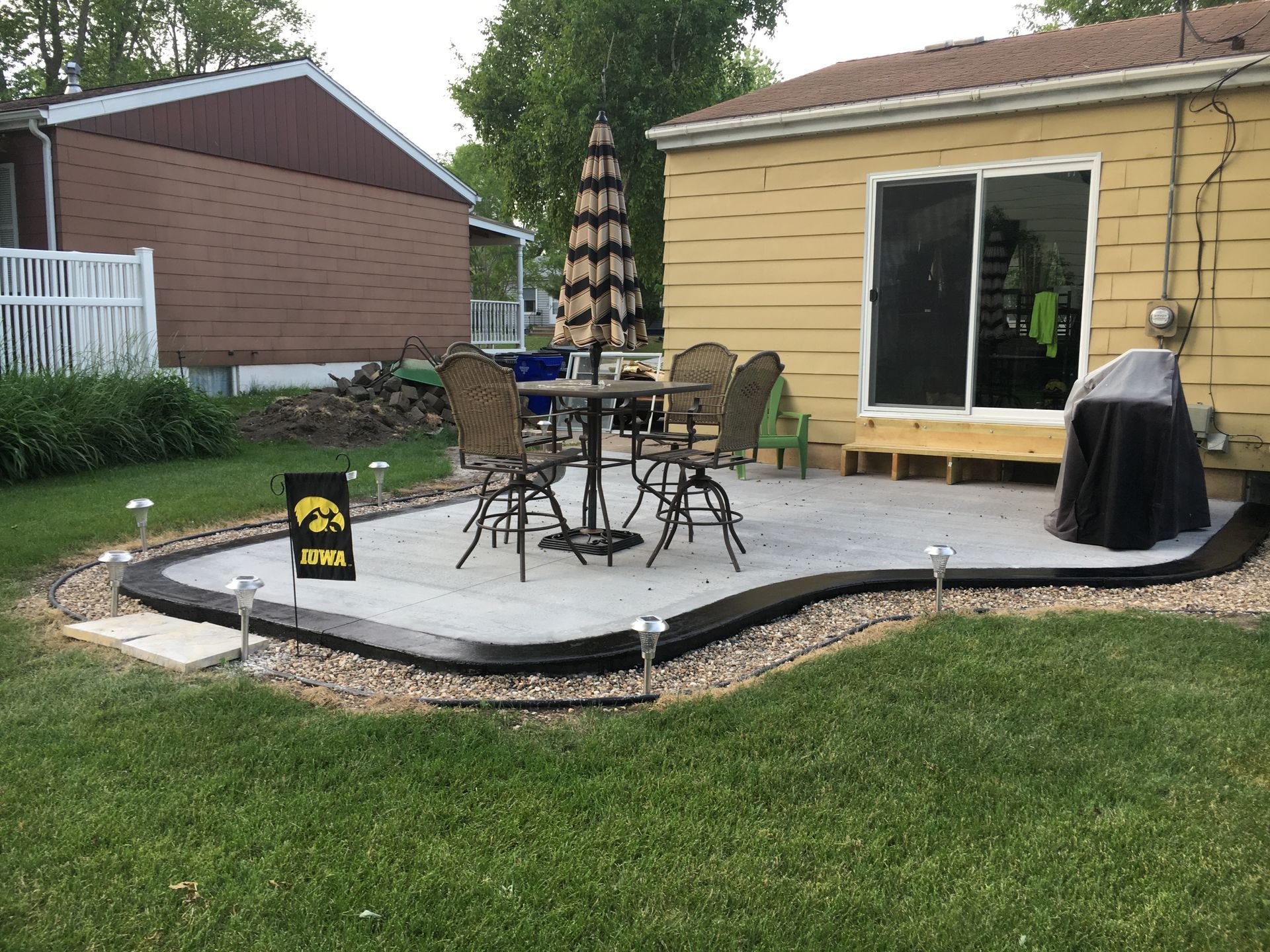 Patio with table, chairs, and umbrella next to a yellow house, surrounded by a rock border, lawn, and solar lights.