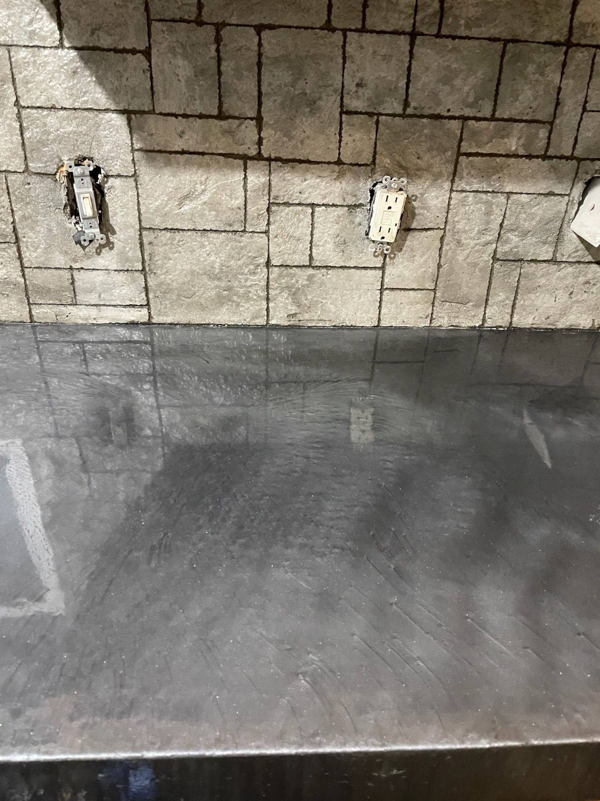 A dark, shiny countertop below a textured, grey brick wall with visible electrical outlets.