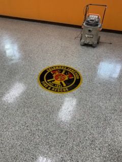 Floor emblem for Kalamara Fire & Rescue, surrounded by grey speckled floor, vacuum cleaner nearby.
