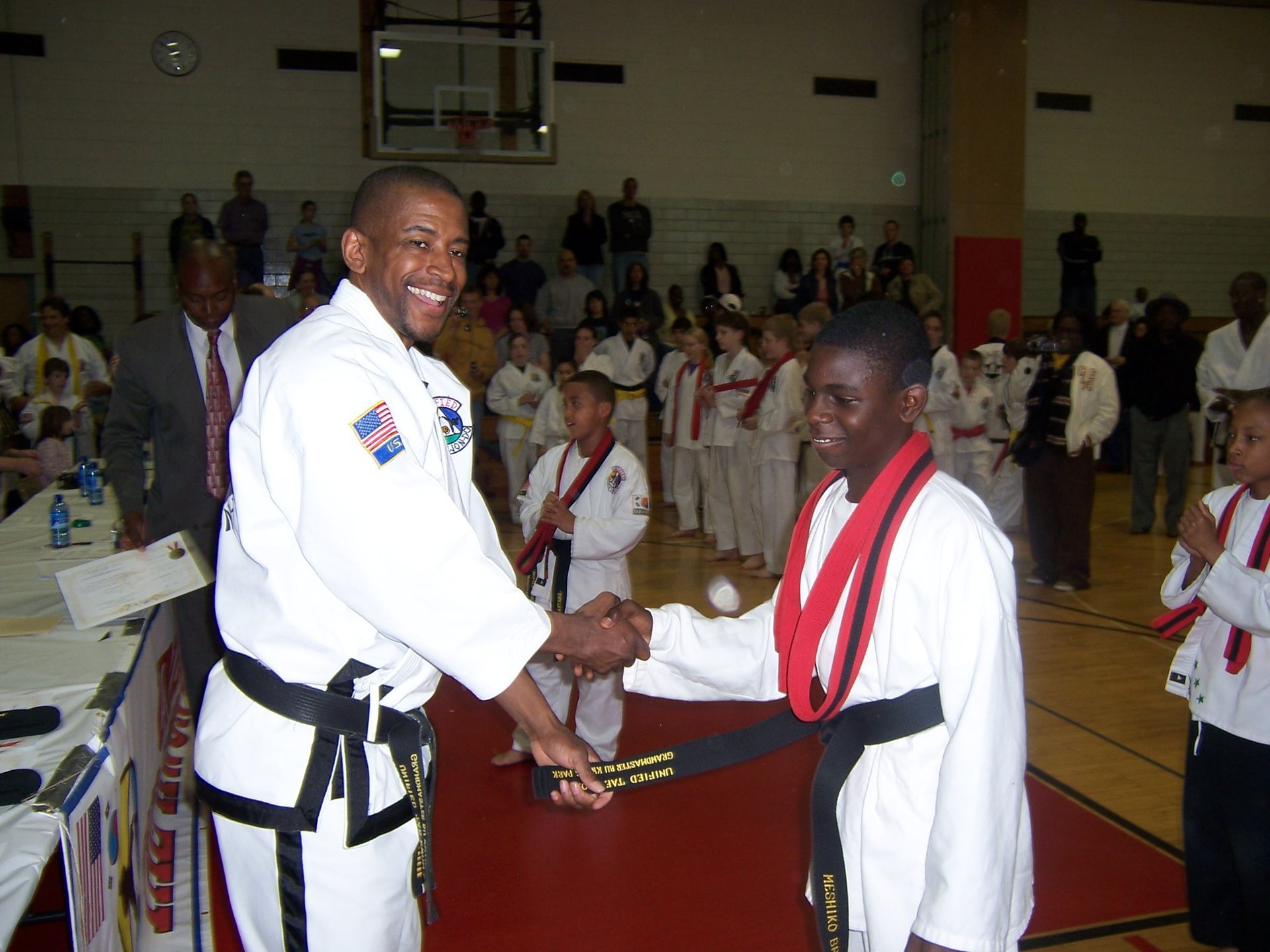 Two martial artists in white uniforms shake hands on a gym floor, with spectators in the background.