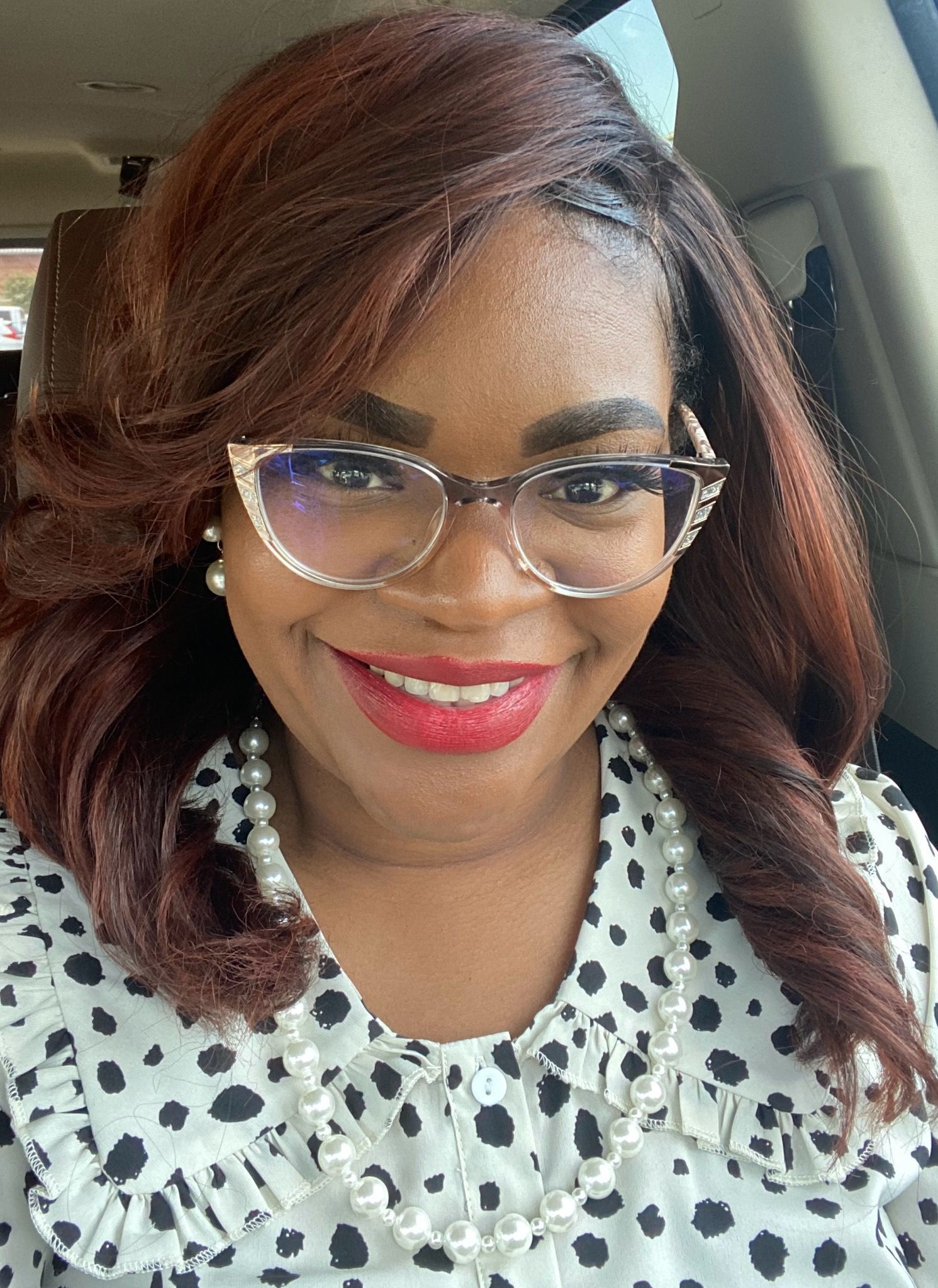 A woman wearing glasses and a pearl necklace is smiling while sitting in a car.