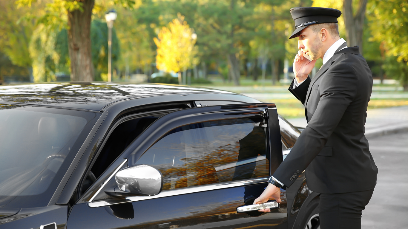 A chauffeur in a black uniform opens a car door while talking on the phone; outdoors.
