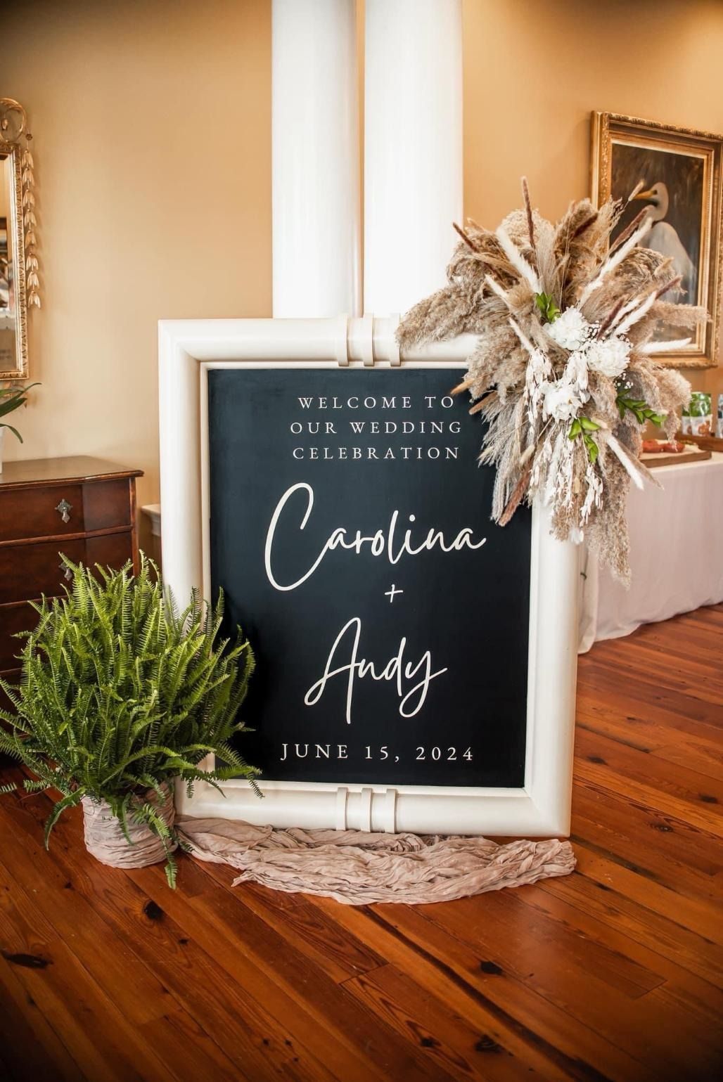 Black wedding welcome sign with white script, greenery, and floral arrangement in a rustic indoor setting