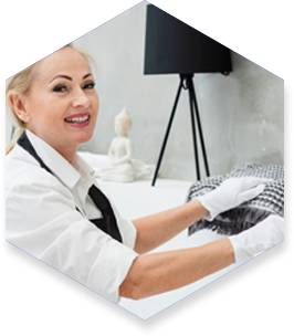 Woman with gloves arranges linens in a minimalist setting; a white Buddha statue and tripod lamp are visible.