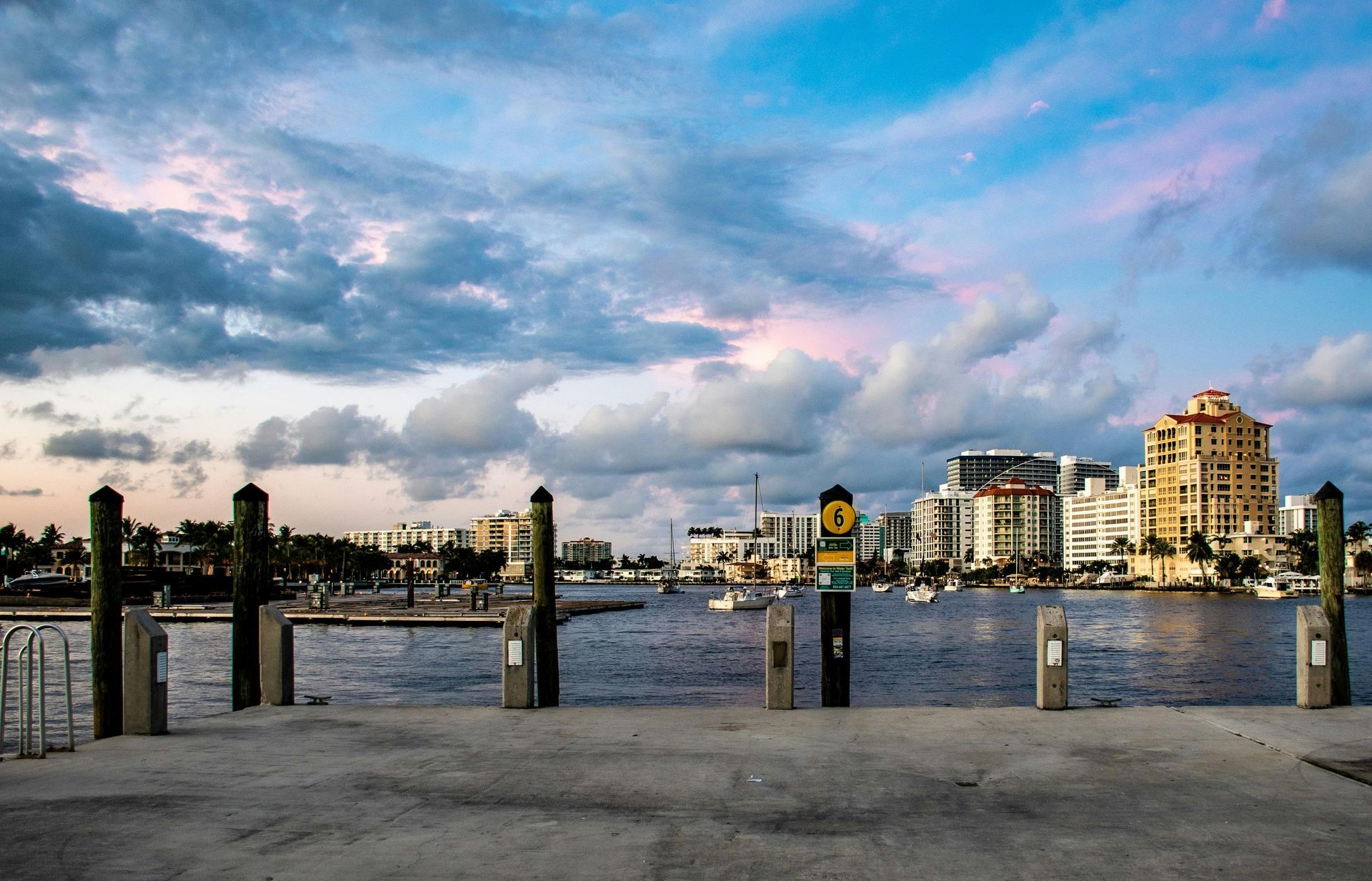Waterfront view at sunset with buildings and boats in the background. The sky has pink and blue hues.