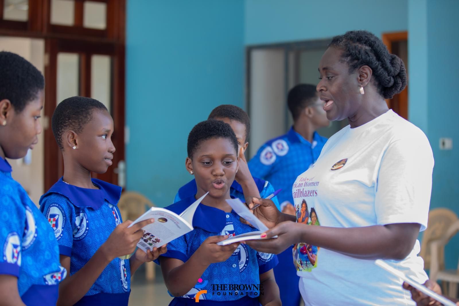 Woman giving books to schoolgirls in blue uniforms. Inside a light blue room.