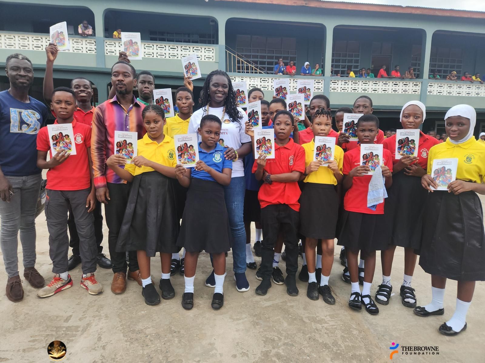 Group of schoolchildren with books, smiling, in front of a building. Some are holding books up.