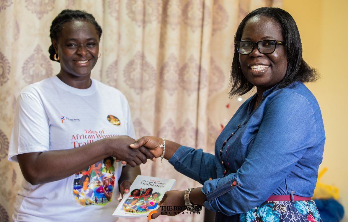 Two women shaking hands indoors; one presents a booklet.