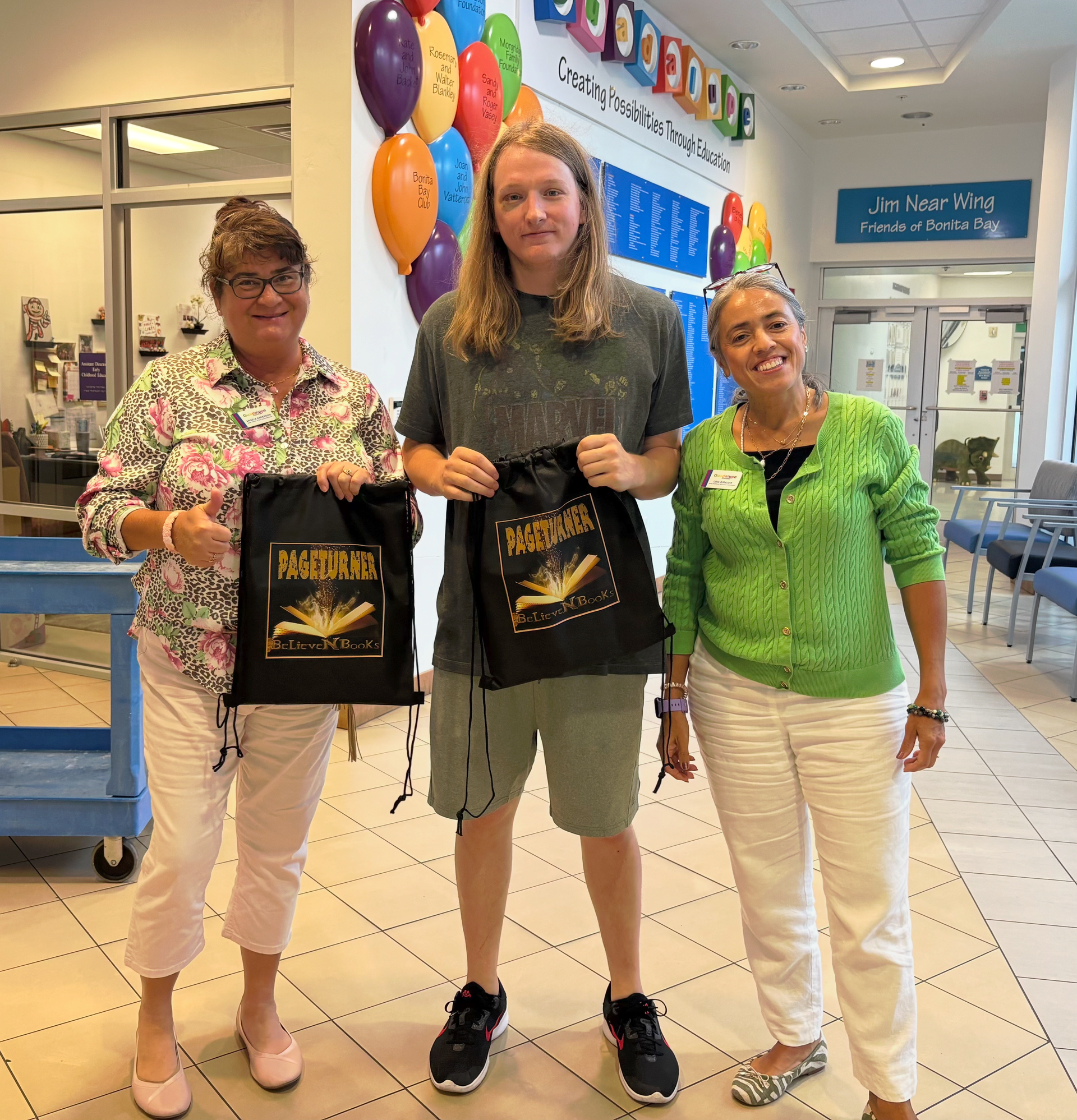 Three people holding tote bags in a brightly lit hallway. Balloons and a sign are in the background.