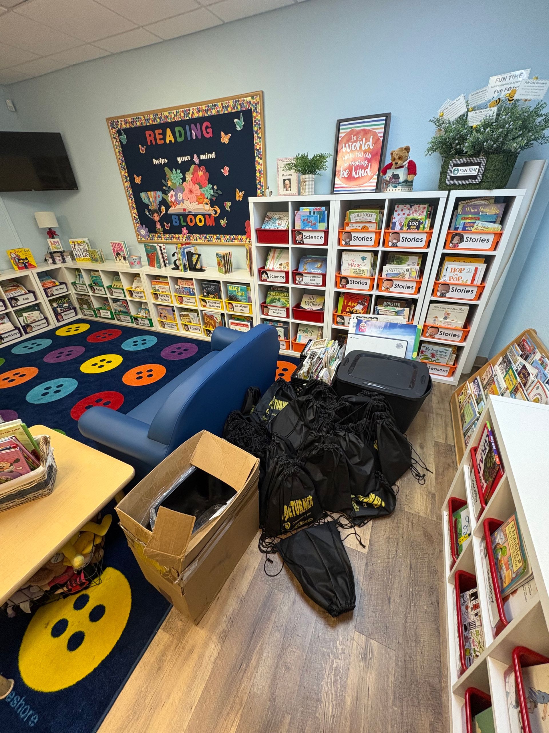 A colorful classroom with bookshelves, reading rug, and a pile of black bags and boxes.