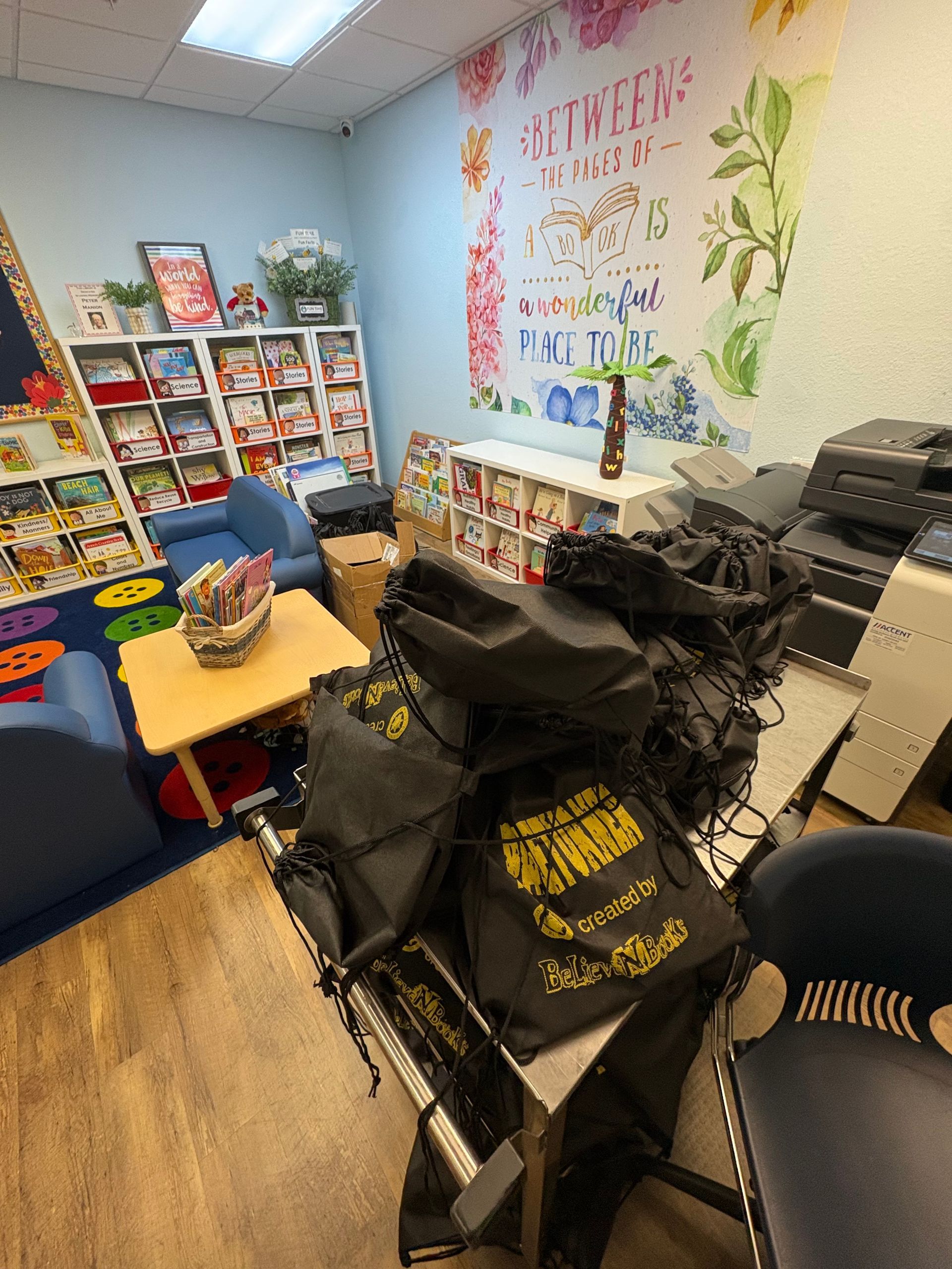Interior of a children's room with bookshelves, blue chairs, small table, and a stack of black bags.