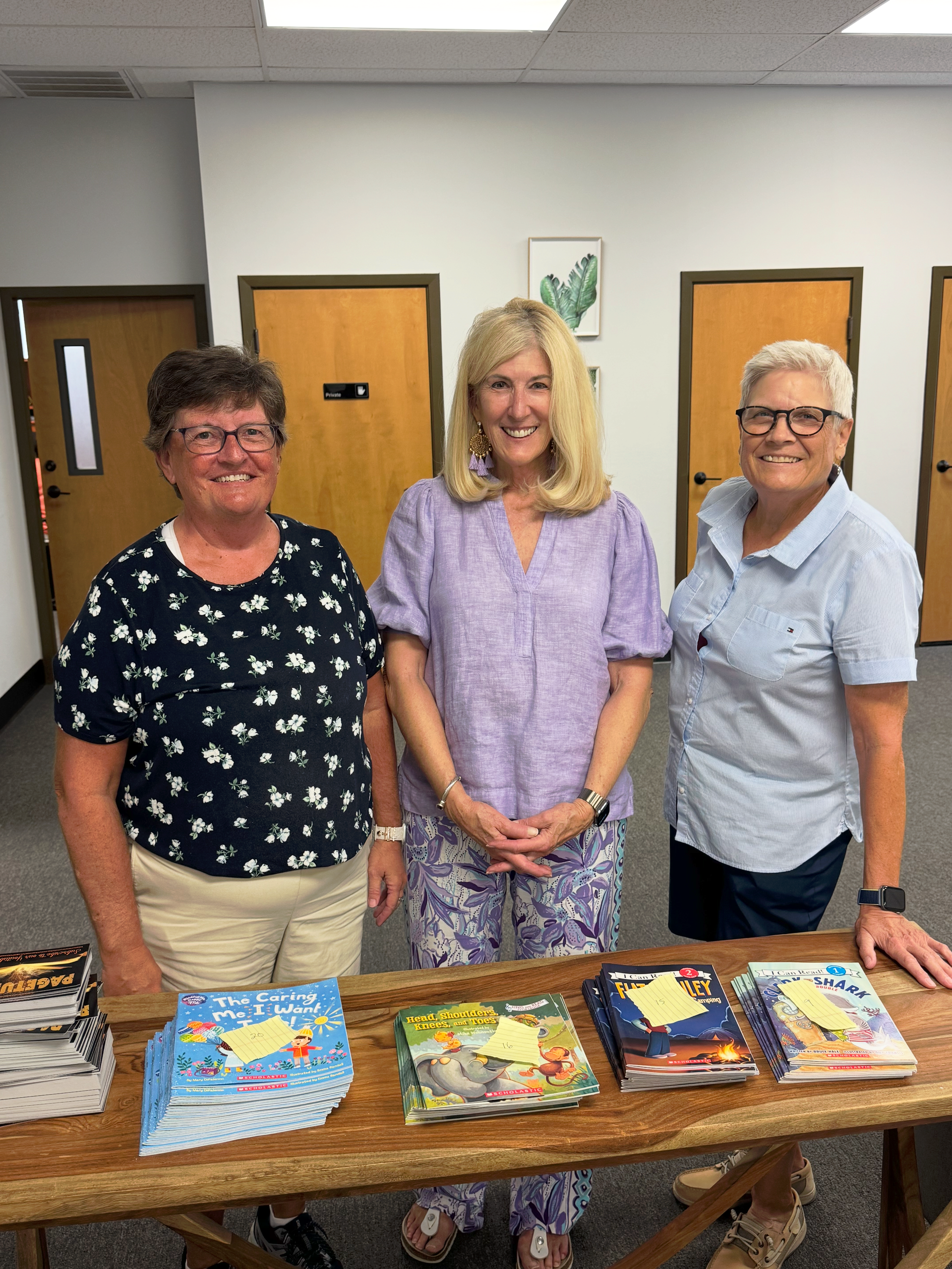 Three women smiling behind a table with books, possibly in a library.