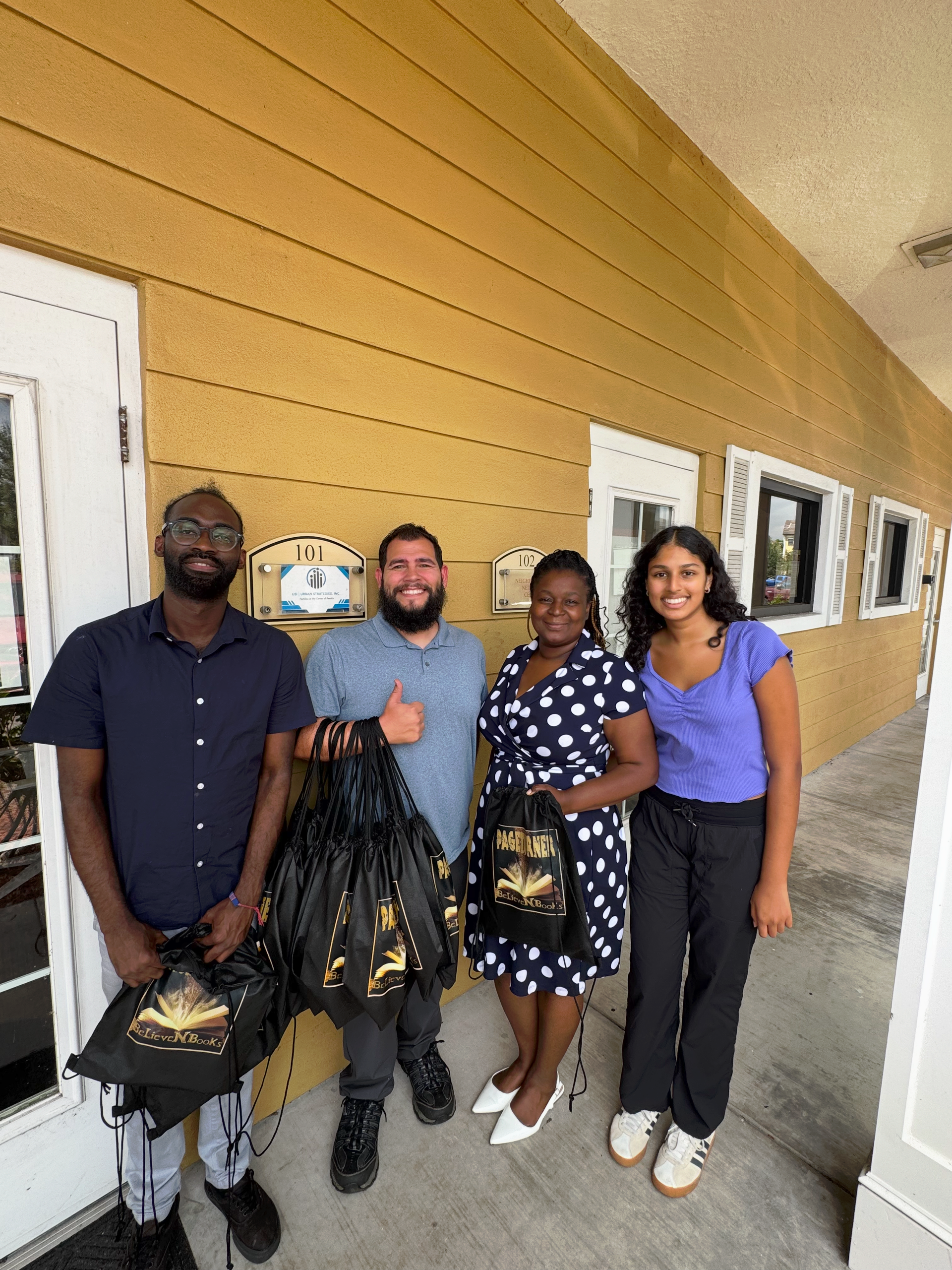 Four people smiling, holding gift bags, posing by a yellow building with a door and windows.