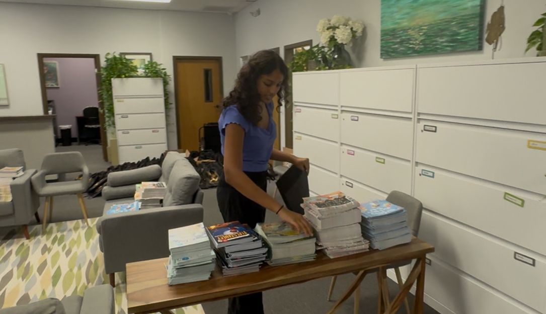 Woman sorts through stacks of papers on a table near lockers in a light-filled office.