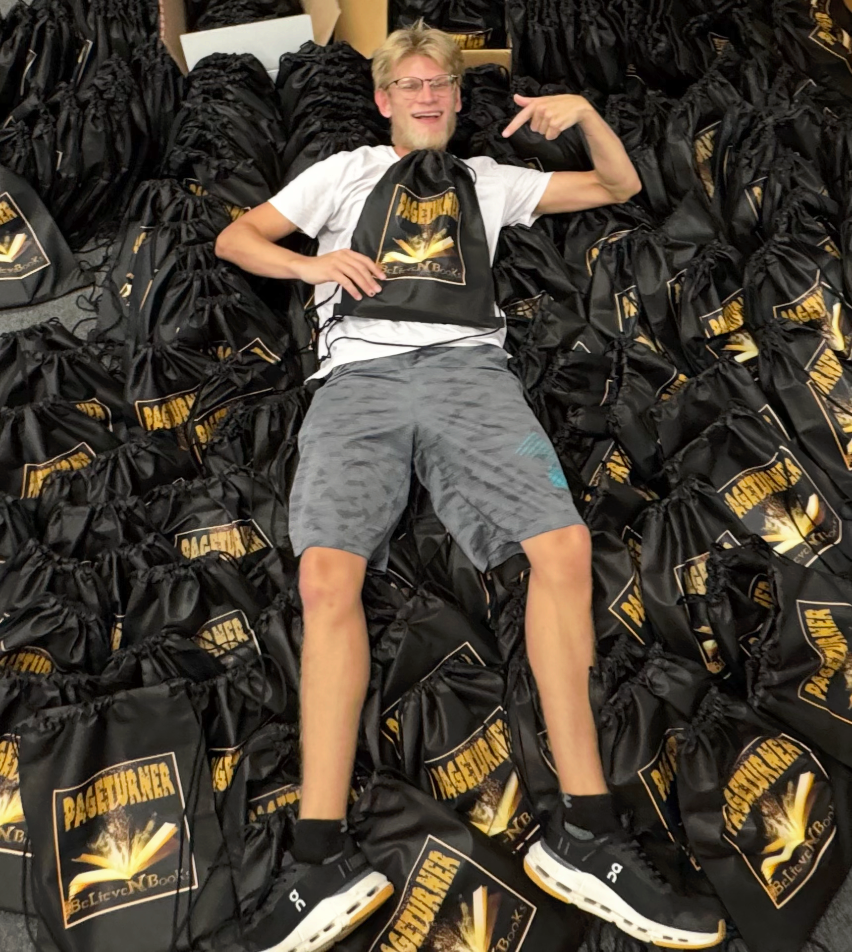 Blond man on black drawstring bags with gold logos; pointing. White shirt, gray shorts. Indoor setting.