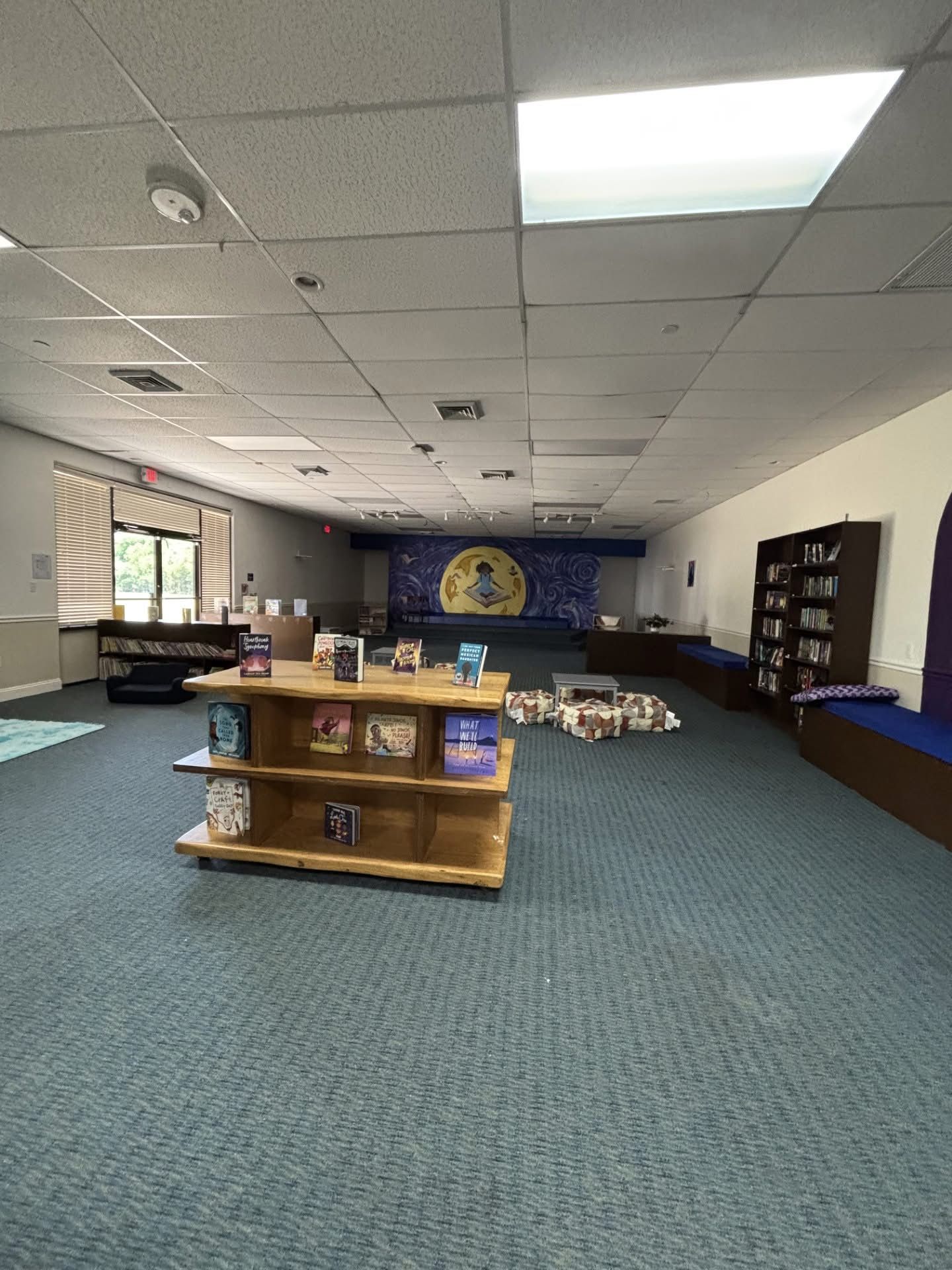 A library interior with a book display, seating, and a mural backdrop. Turquoise carpet and shelves.