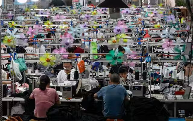 Workers at sewing machines in a factory, surrounded by colorful fans, likely in a hot environment.