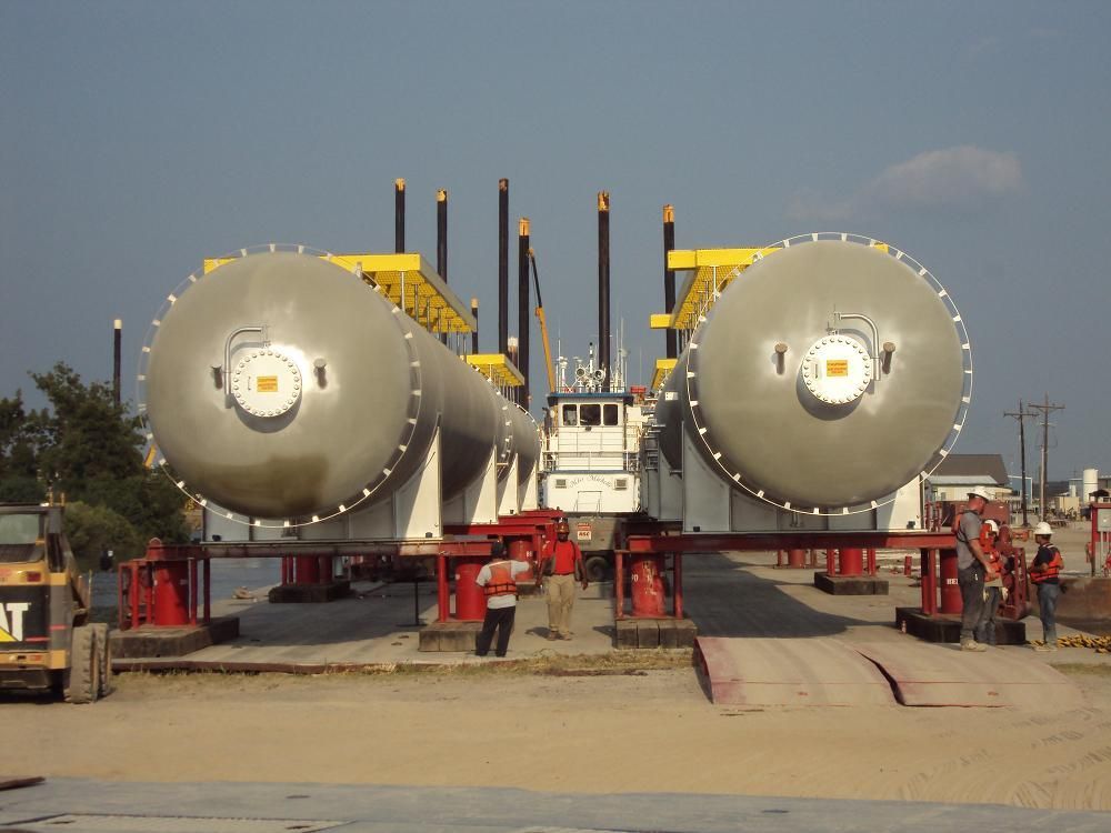 Large, gray cylindrical tank on a flatbed truck, traveling on a road. Banner on tank.