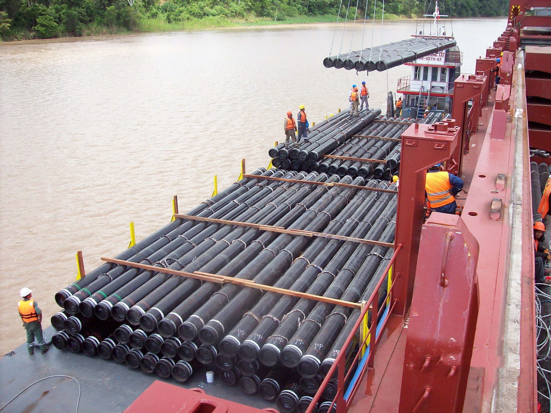 Pipes being loaded onto a cargo ship on a river, workers in orange vests.